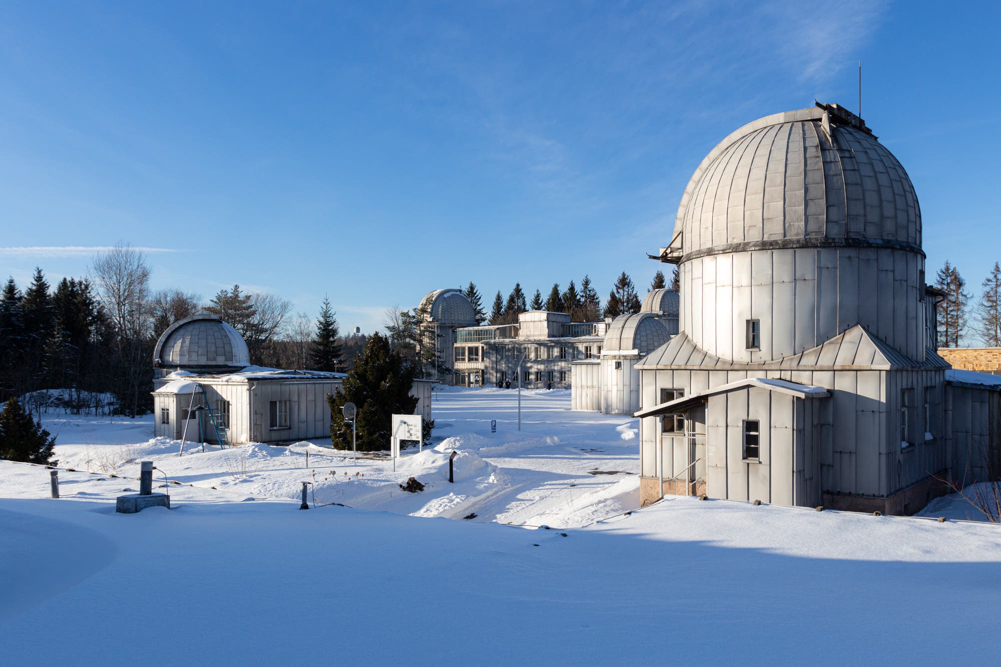 Eine verschneite Landschaft mit mehreren Observatorien der Sternwarte Sonneberg, die durch ihre charakteristischen Kuppeln erkennbar sind. Die Gebäude sind von schneebedeckten Flächen und Bäumen umgeben. Der Himmel ist klar und blau. Es ist ein heller sonniger Wintertag.
