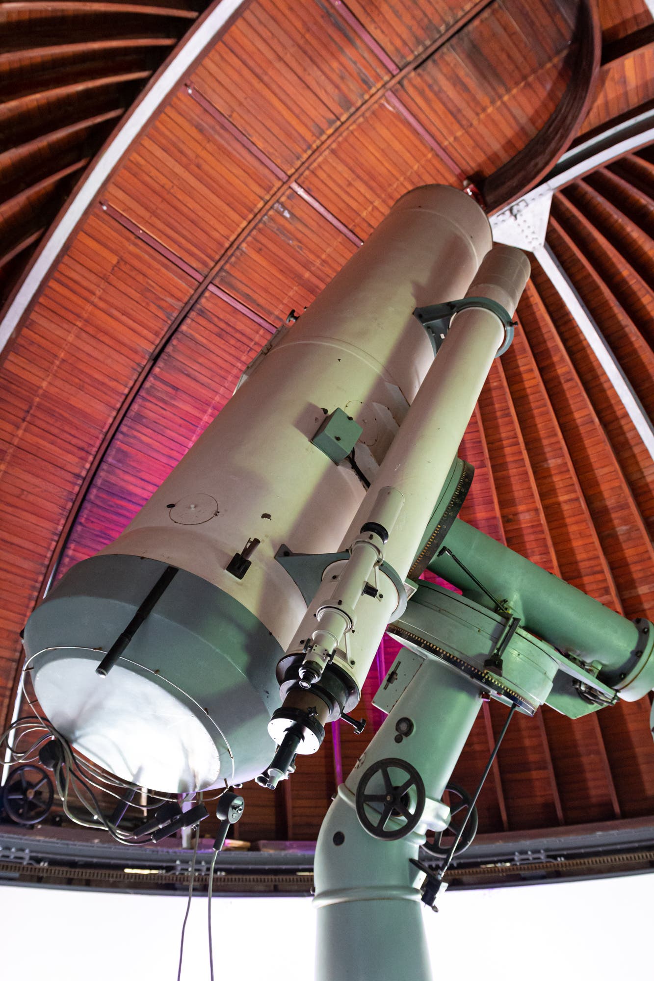 Ein großes Teleskop in der Sternwarte Sonneberg ist nach oben gerichtet. Die Struktur besteht aus mehreren zylindrischen Komponenten und ist in einem hölzernen Kuppeldach untergebracht. Das Teleskop ist mit verschiedenen mechanischen Teilen und Kabeln ausgestattet. Die Umgebung ist gut beleuchtet, was die Details der Konstruktion hervorhebt.