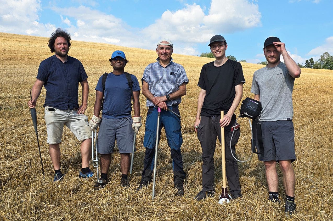 Fünf Personen stehen auf einem abgeernteten Feld unter einem blauen Himmel mit weißen Wolken. Sie tragen Freizeitkleidung und halten verschiedene Werkzeuge, die auf landwirtschaftliche oder geologische Arbeiten hindeuten. Die Gruppe lächelt in die Kamera, während sie in einer Linie posiert. Im Hintergrund erstreckt sich das goldene Feld bis zum Horizont.