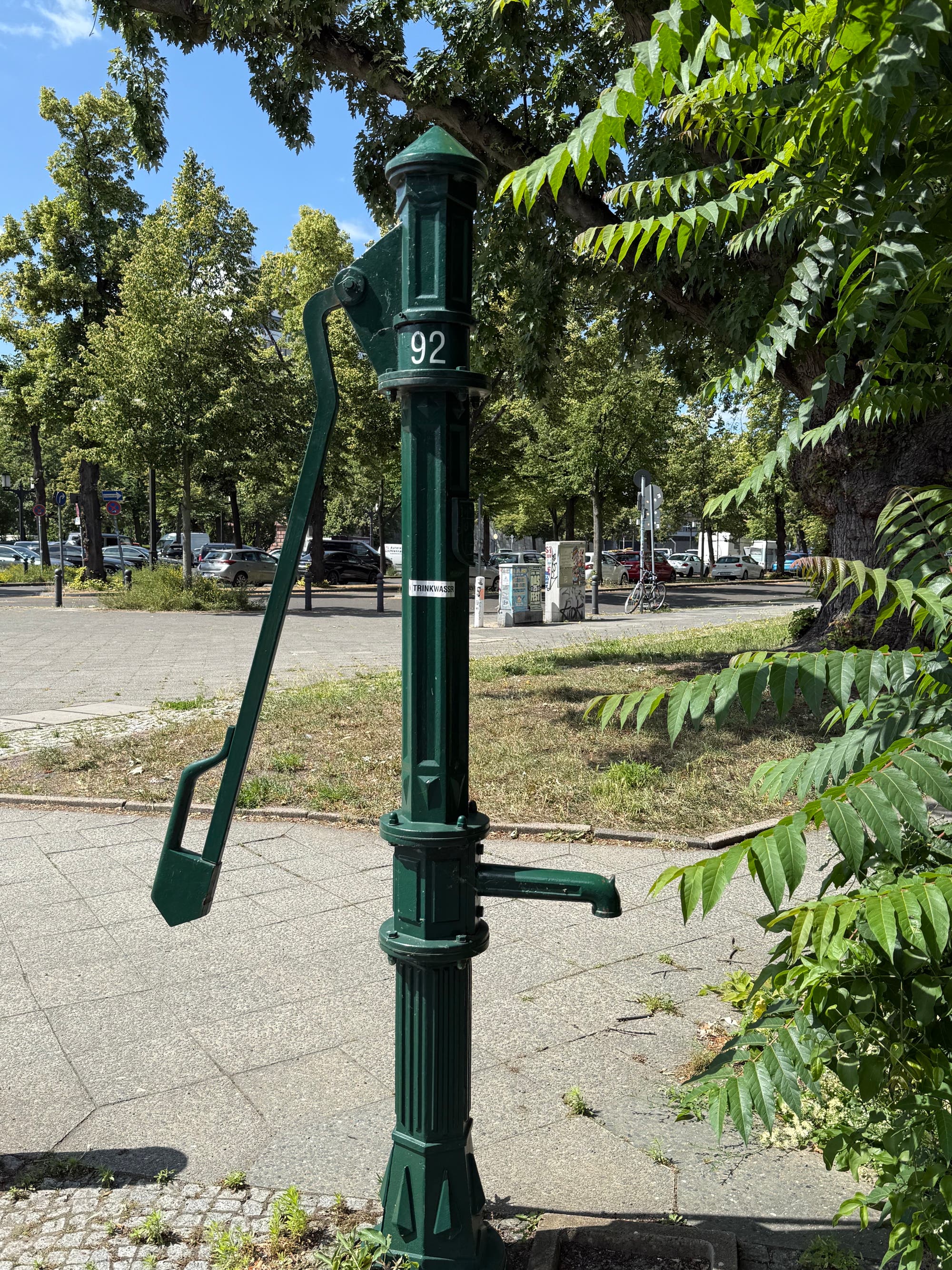 Eine grüne Wasserpumpe steht auf einem Gehweg, umgeben von Bäumen und Pflanzen. Die Pumpe hat die Nummer "92" auf der Seite. Im Hintergrund sind geparkte Autos und weitere Bäume zu sehen. Die Szene ist bei Tageslicht aufgenommen, und das Wetter scheint klar zu sein.