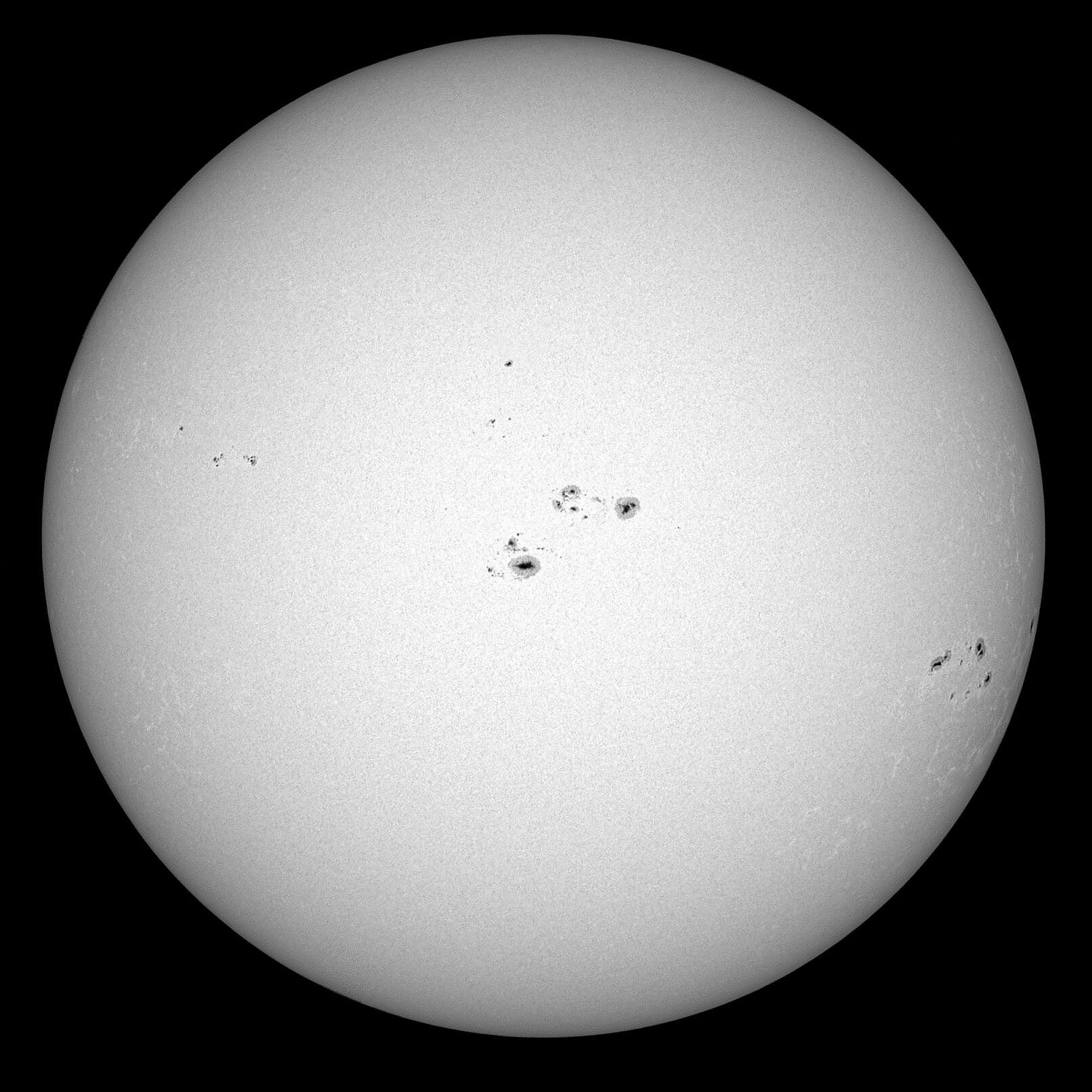 Una imagen en blanco y negro del sol, que muestra varias manchas solares oscuras en su superficie. Estas manchas solares están distribuidas irregularmente y varían en tamaño. La figura ilustra la actividad en la superficie del sol y se utiliza para observar y analizar fenómenos solares. 