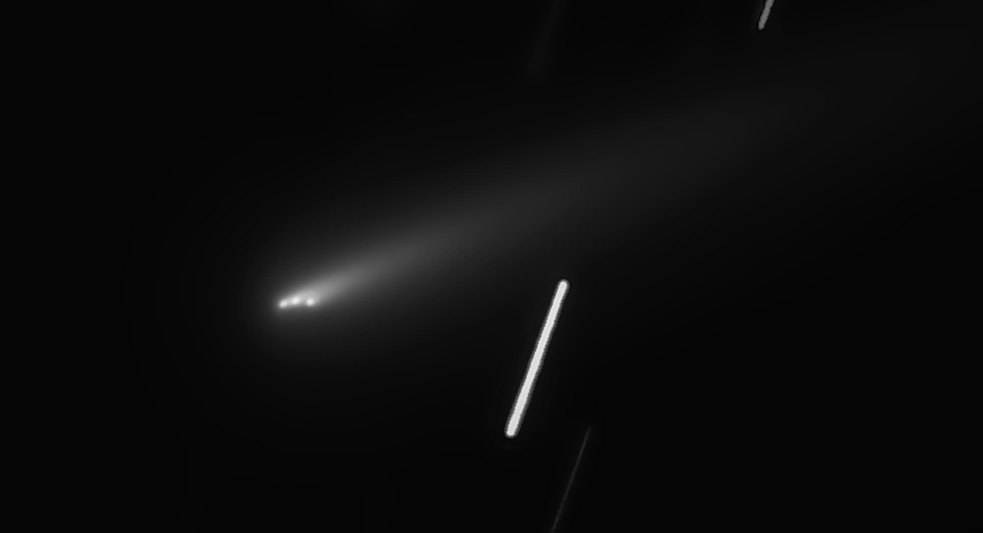 Ein astronomisches Bild zeigt einen Kometen mit einem leuchtenden Kern und einem langen, diffusen Schweif, der sich nach rechts erstreckt. Der Hintergrund ist schwarz, was den Kometen hervorhebt. Mehrere helle, diagonale Linien sind sichtbar, die Sterne darstellen, die durch die Bewegung der Kamera w&auml;hrend der Aufnahme entstanden sind.