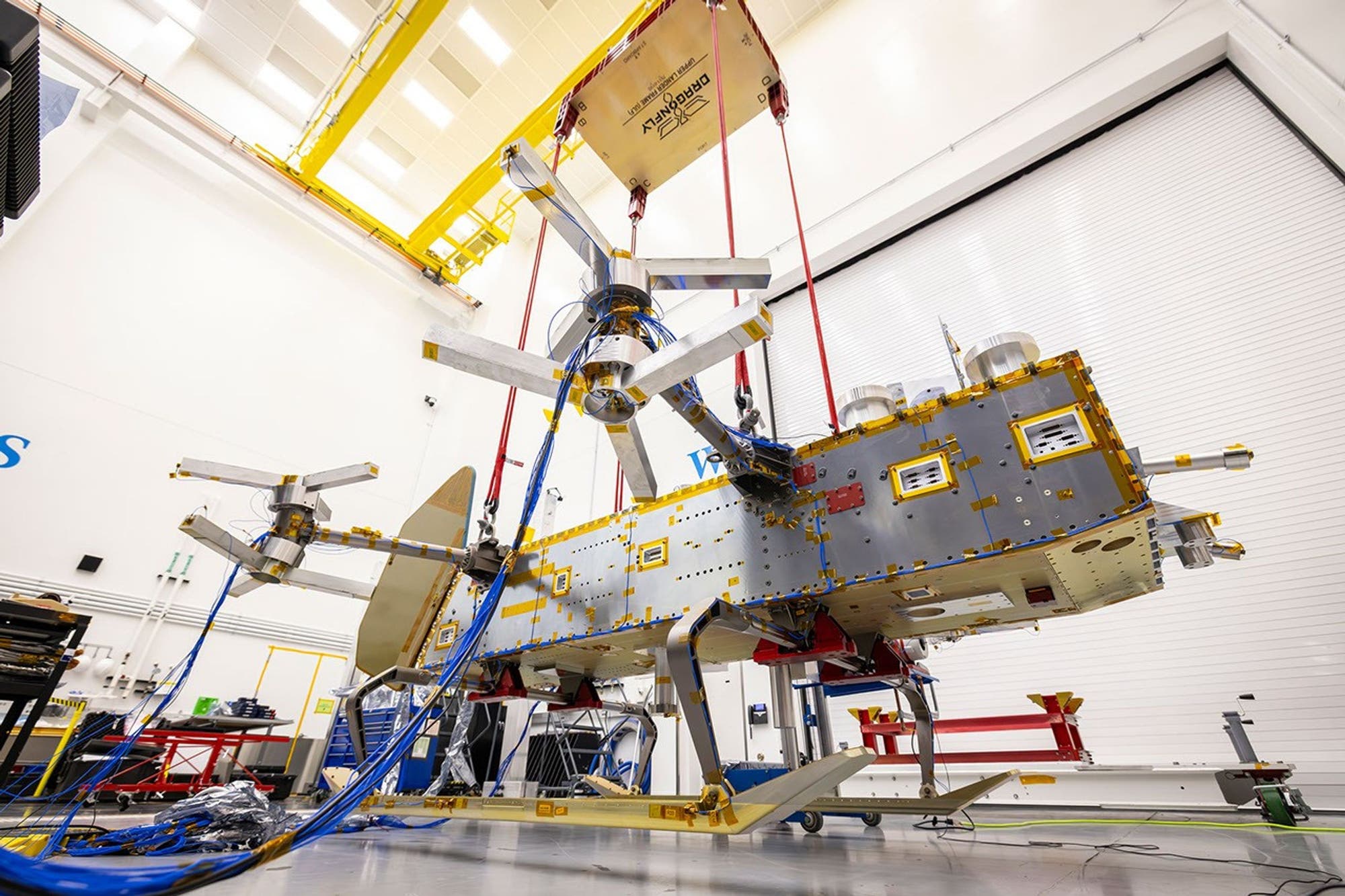 Ein Satellit wird in einer großen, hell erleuchteten Werkshalle montiert. Der Satellit ist an mehreren roten Gurten aufgehängt und zeigt komplexe mechanische Strukturen und Verkabelungen. Im Hintergrund sind Werkzeuge und Ausrüstungen sichtbar. Die Umgebung wirkt industriell und technisch. Ein Satellit wird in einer großen, hell erleuchteten Werkshalle montiert. Der Satellit ist an mehreren roten Gurten aufgehängt und zeigt komplexe mechanische Strukturen und Verkabelungen. Im Hintergrund sind Werkzeuge und Ausrüstungen sichtbar. Die Umgebung wirkt industriell und technisch.