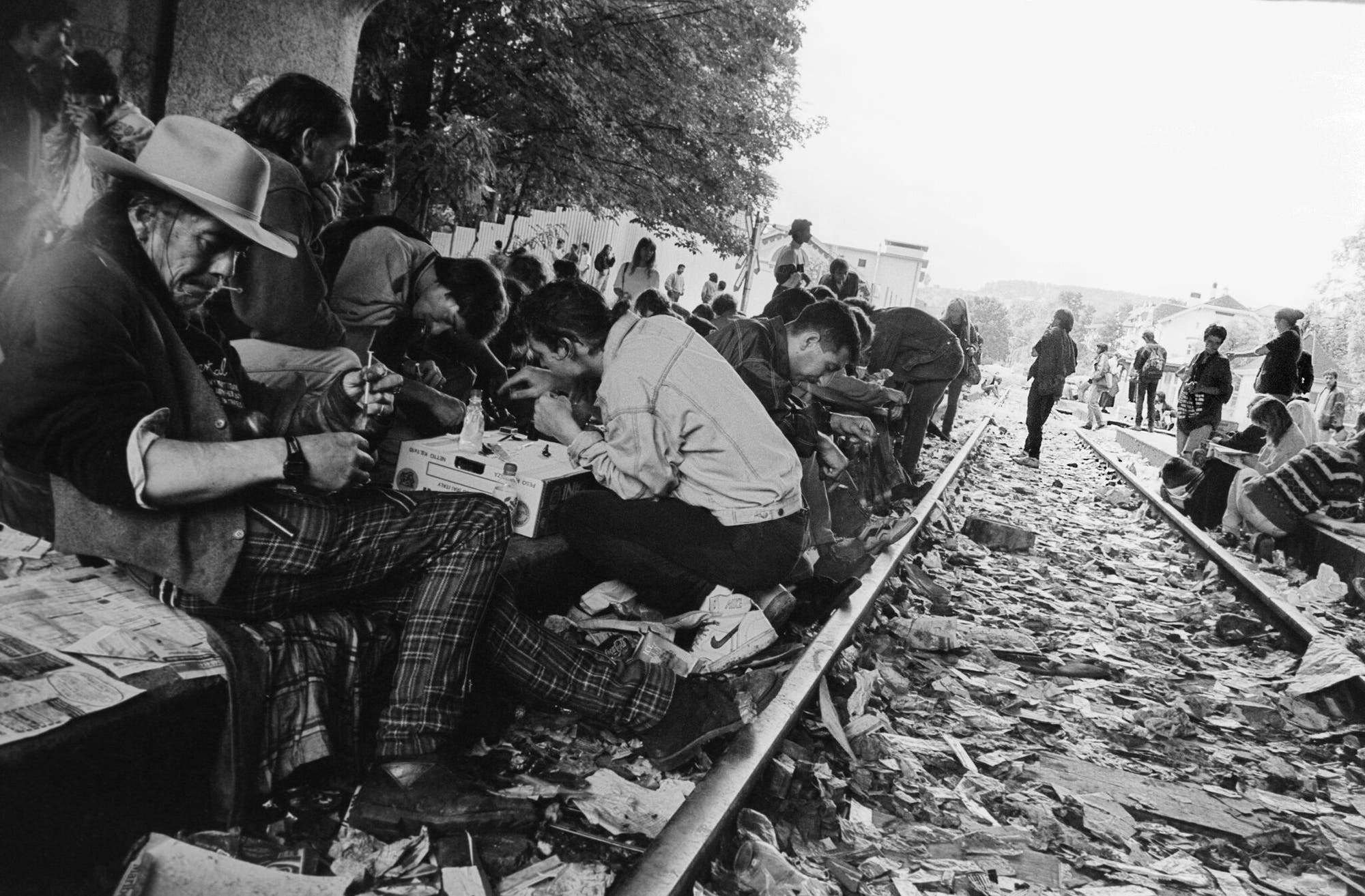 Eine Gruppe von Menschen sitzt und steht auf Bahngleisen, umgeben von verstreutem Müll. Einige Personen sind in Gespräche vertieft oder konsumieren Drogen. Im Vordergrund sitzt ein Mann mit Hut, der eine Spritze in den Händen hält. Die Szene wirkt chaotisch und zeigt eine Ansammlung von Menschen in einer urbanen Umgebung. Bäume und Gebäude sind im Hintergrund sichtbar.