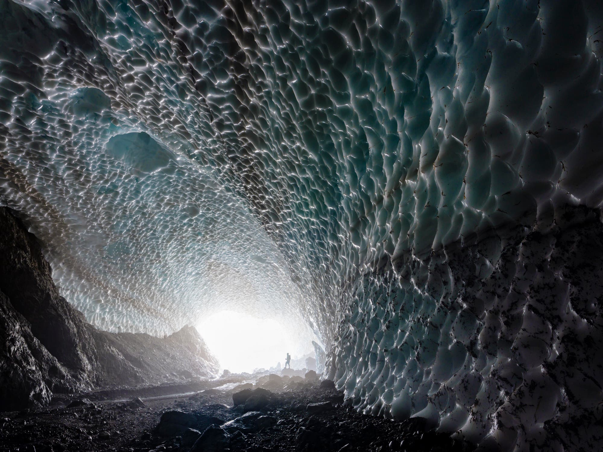 Eine Person steht am Ende einer großen Eishöhle, die von unregelmäßigen, wellenförmigen Eisstrukturen bedeckt ist. Das Licht strömt von einem hellen Ausgang am Ende der Höhle herein, wodurch die Textur des Eises hervorgehoben wird. Die Szene vermittelt ein Gefühl von Größe und Kälte. Im Vordergrund sind dunkle Felsen zu sehen.