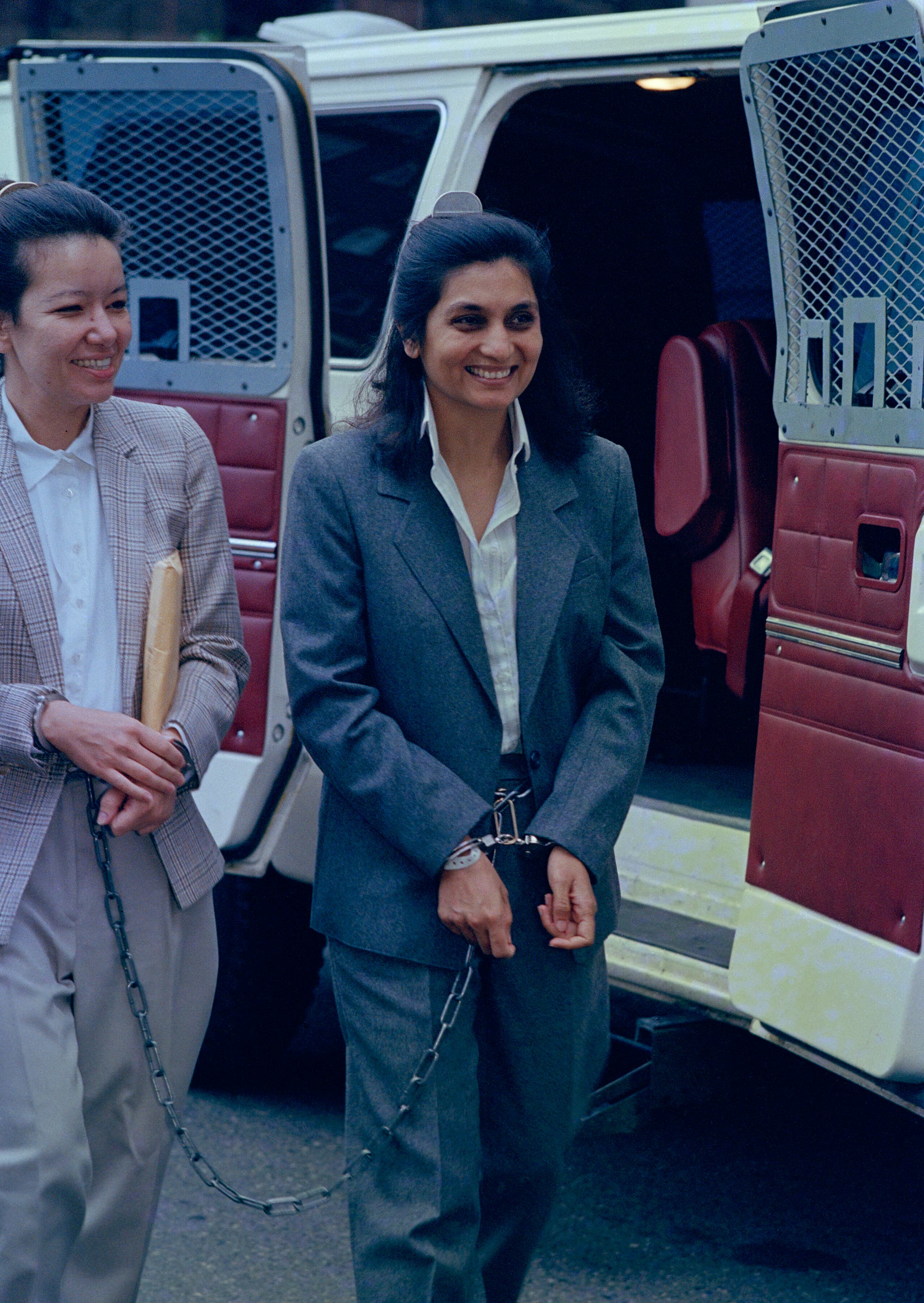 Zwei Personen in Anzügen stehen vor einem Fahrzeug mit offenen Türen. Beide sind mit Handschellen gefesselt und durch eine Kette verbunden. Sie lächeln und scheinen in einem formellen Kontext zu stehen. Im Hintergrund ist ein Fahrzeug mit Gittertüren zu sehen.