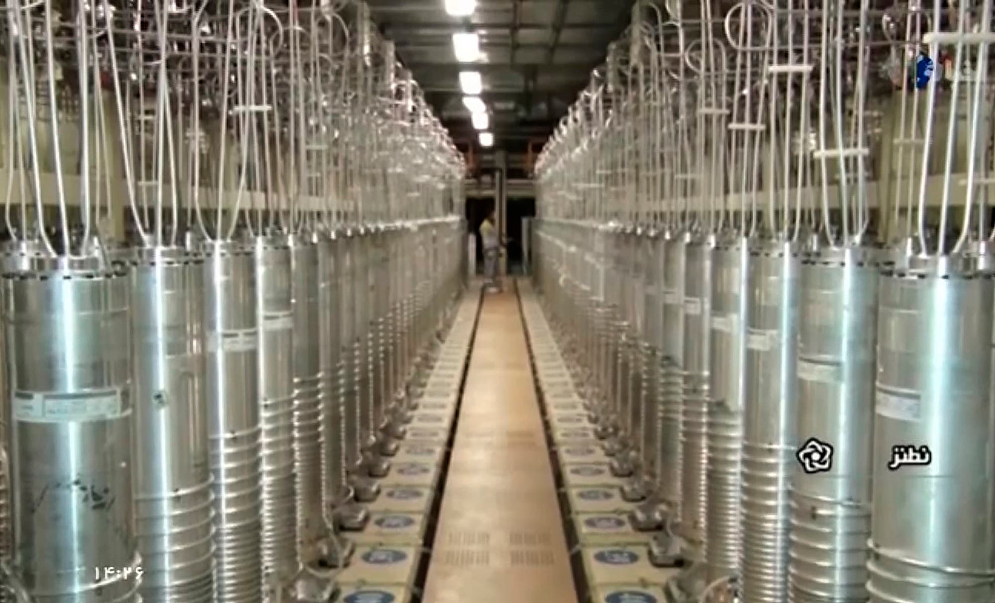 Eine lange Reihe von metallischen Zentrifugen in einem industriellen Raum, die für die Urananreicherung verwendet werden. Die Zentrifugen sind in zwei parallelen Reihen angeordnet, die einen schmalen Gang dazwischen bilden. An den Zentrifugen sind Kabel und Schläuche befestigt. Der Raum ist gut beleuchtet und hat eine hohe Decke. Auf der rechten Seite des Bildes sind arabische Schriftzeichen zu sehen. Keine Personen sind erkennbar.