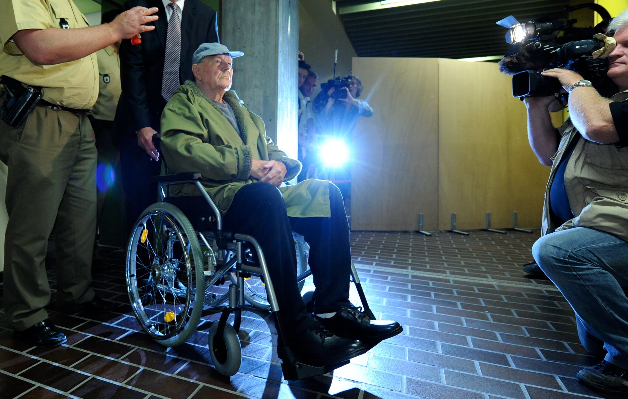 Ein alter Mann sitzt in einem Rollstuhl, umgeben von mehreren Kameraleuten und Sicherheitsbeamten. Die Szene ist in einem Innenraum mit gedämpfter Beleuchtung; ein Kamerablitz leuchtet im Hintergrund auf. Der Mann im Rollstuhl trägt einen grünen Mantel und eine blaue Kappe. Die Kameras sind auf ihn fokussiert. Die Atmosphäre wirkt angespannt.