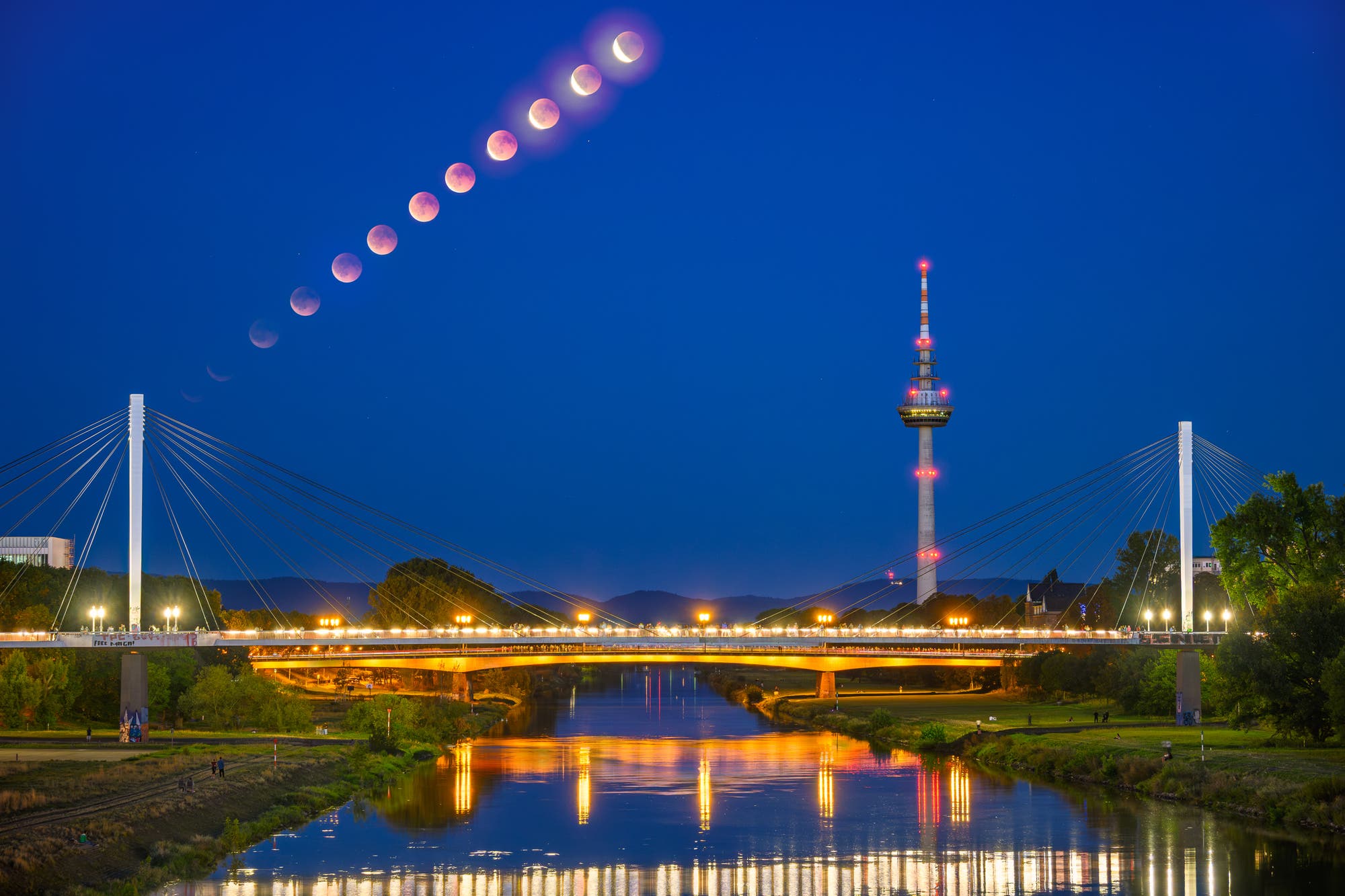 Eine nächtliche Stadtlandschaft mit einer beleuchteten Brücke und einem Fernsehturm im Vordergrund. Am Himmel ist eine Abfolge von Mondphasen während einer Mondfinsternis zu sehen, die von links nach rechts verläuft. Der Fluss unter der Brücke spiegelt die Lichter wider, und der Himmel ist klar und dunkelblau. Schlüsselwörter: Mondfinsternis, Brücke, Fernsehturm, Nacht, Reflexion.
