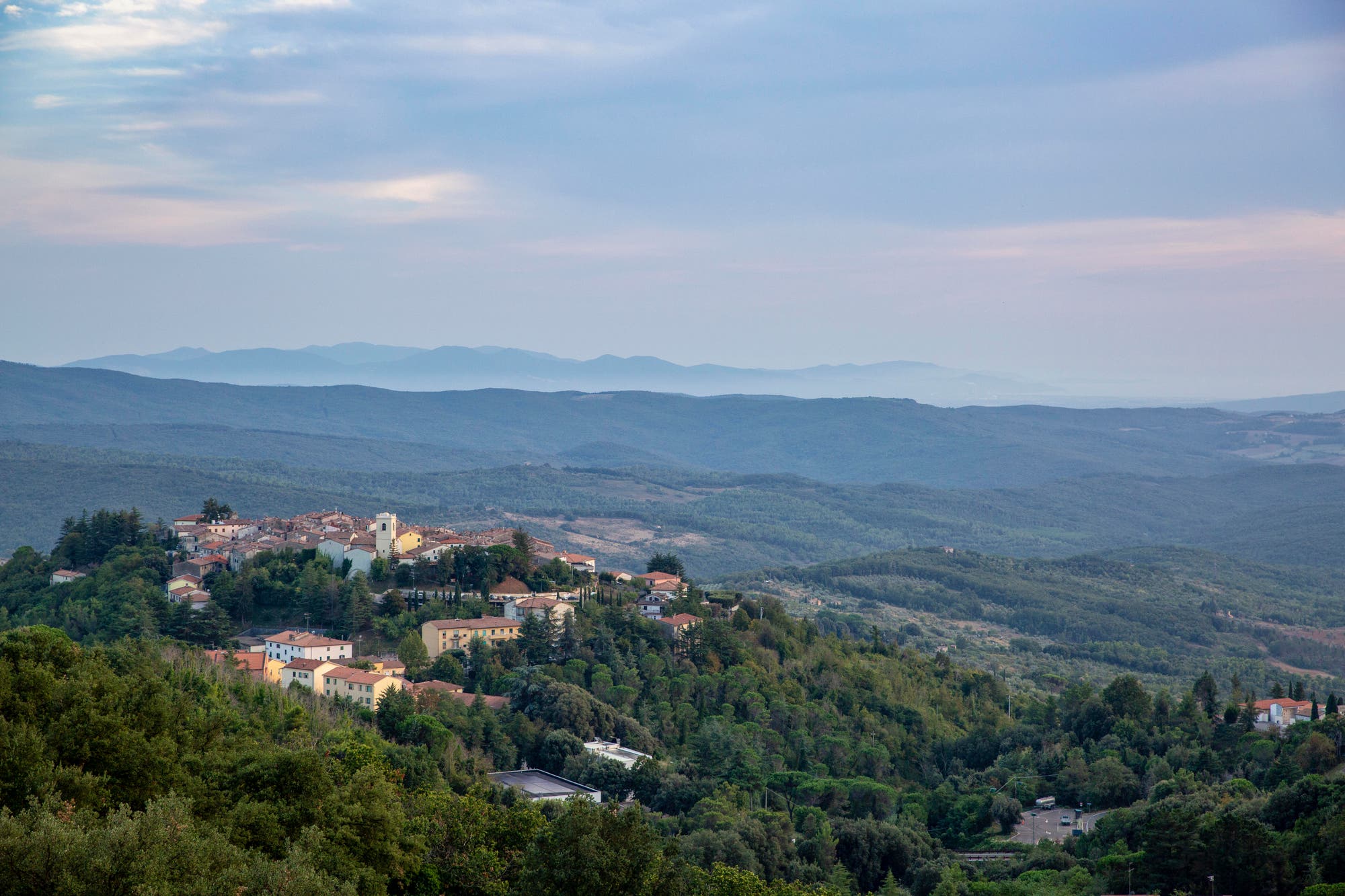Eine malerische Aussicht auf ein kleines Dorf, das auf einem bewaldeten Hügel liegt, umgeben von ausgedehnten grünen Wäldern und Hügelketten, die sich bis zum Horizont erstrecken. Der Himmel ist leicht bewölkt, was eine ruhige und friedliche Atmosphäre schafft. Im Dorf sind mehrere Gebäude mit roten Dächern und eine Kirche mit einem Glockenturm zu sehen.