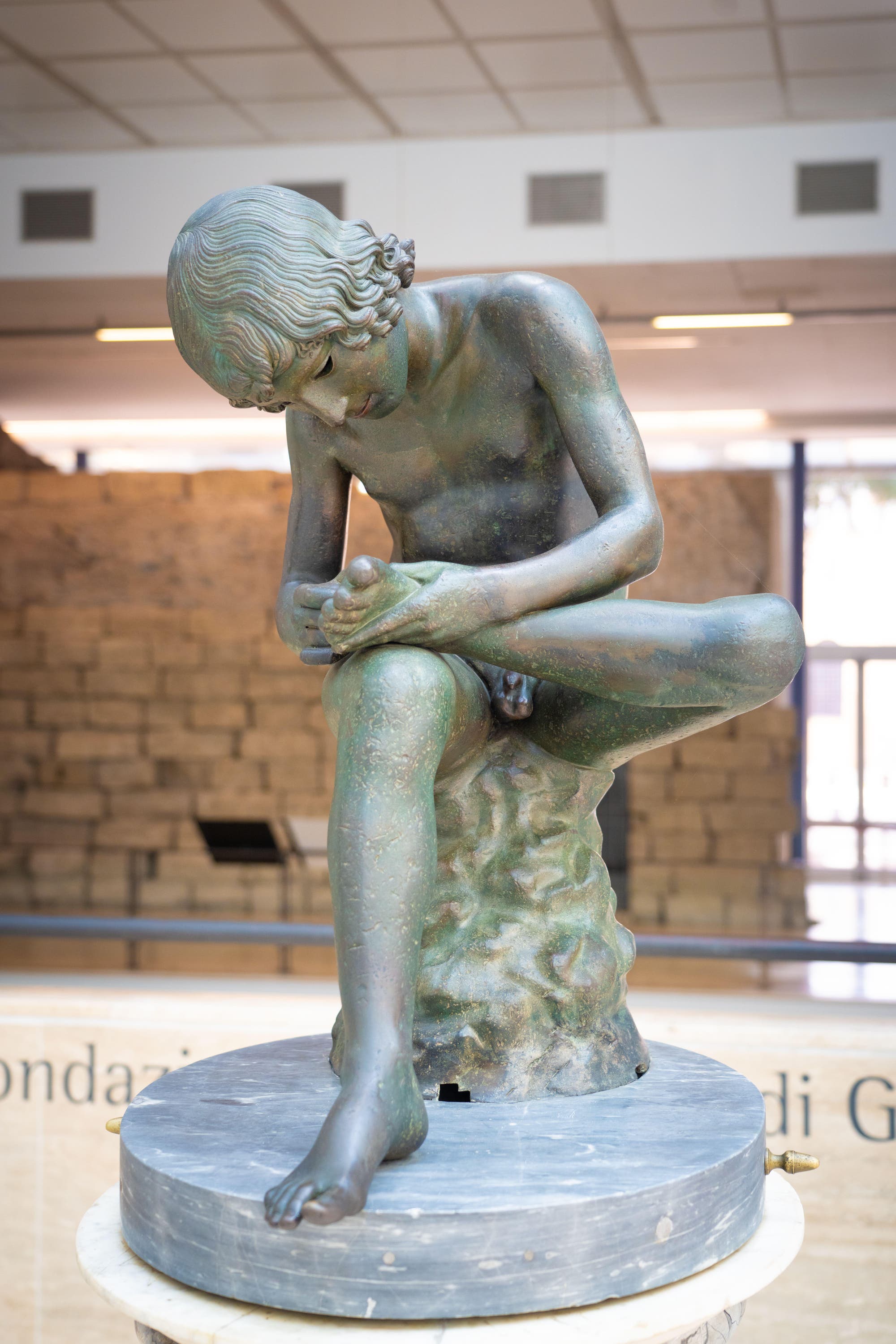 Eine Bronzestatue eines jungen Mannes, der auf einem Felsen sitzt und einen Dorn aus seinem Fuß zieht. Die Skulptur befindet sich in einem Museum mit Steinwänden und einer großen Fensterfront im Hintergrund. Der Raum ist gut beleuchtet und die Statue steht auf einem runden Sockel.