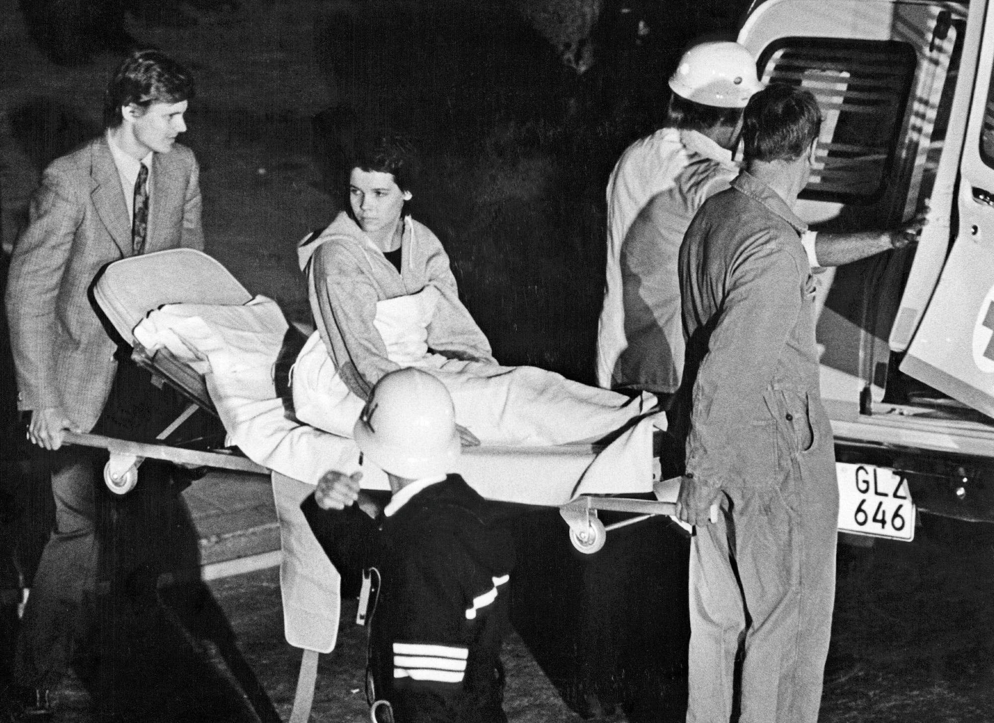 S/W-Aufnahme: Junge Frau wird auf einer Trage zum Krankenwagen gebracht, sie schaut zurück in die Richtung, aus der sie gekommen sind.