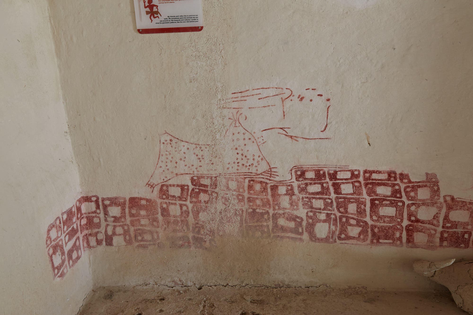 Zu sehen ist die Rekonstruktion einer Wandmalerei in Çatalhöyük. Es zeigt rötliche Gebäude und einen Berg mit doppeltem Gipfel, der Feuer spuckt. Der Hintergrund ist eine beige Wand mit sichtbaren Gebrauchsspuren.