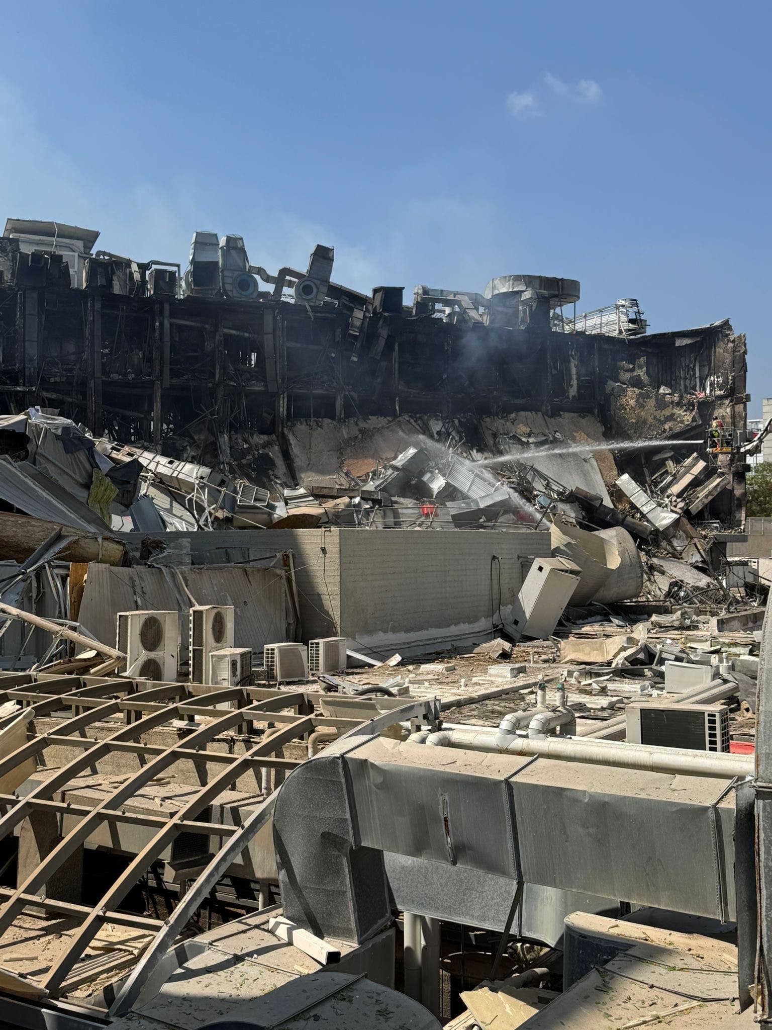Ein zerstörtes Gebäude mit eingestürzten Wänden und Trümmern, das von Rauchschwaden umgeben ist. Im Vordergrund sind beschädigte Klimaanlagen und Metallstrukturen zu sehen. Der Himmel ist klar und blau. Die Szene deutet auf einen kürzlichen Brand oder eine Explosion hin.