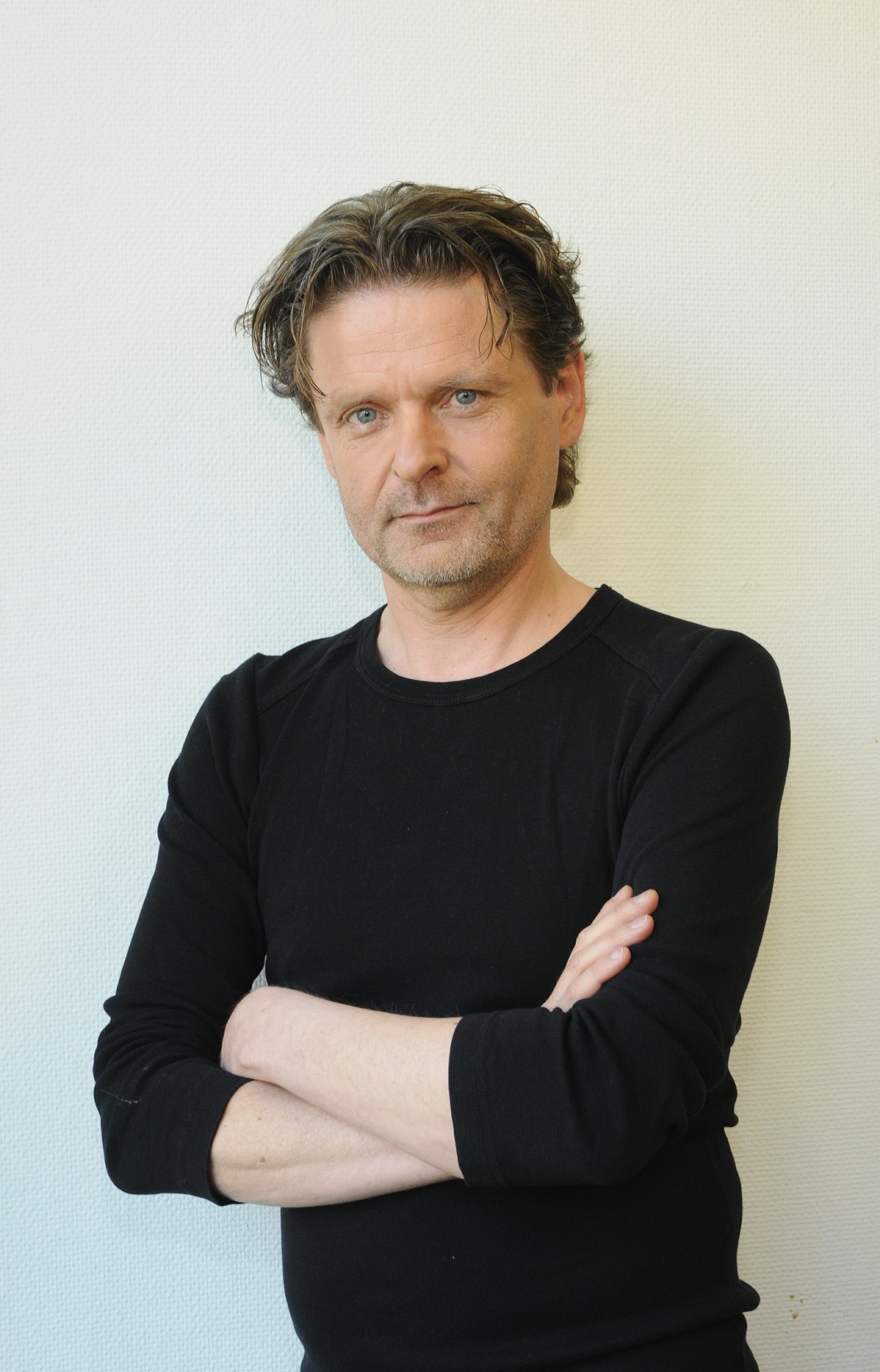 Ein Mann in einem schwarzen Pullover lächelt in die Kamera. Der Hintergrund ist neutral und hell, was den Fokus auf das Gesicht und den Ausdruck des Mannes legt.