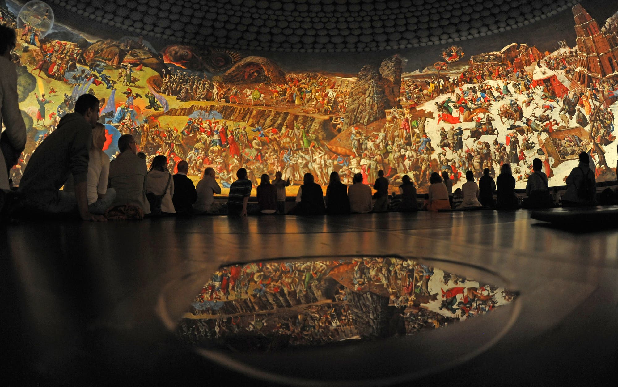 Eine Gruppe von Menschen sitzt in einem dunklen Raum vor einem großen, farbenfrohen Panoramagemälde, das eine historische Schlacht darstellt. Die Szene zeigt viele Figuren in historischer Kleidung, die in verschiedenen Aktionen dargestellt sind. Im Vordergrund spiegelt sich das Gemälde auf einer glänzenden Oberfläche. Die Atmosphäre ist ruhig und konzentriert, während die Betrachter das Kunstwerk betrachten.