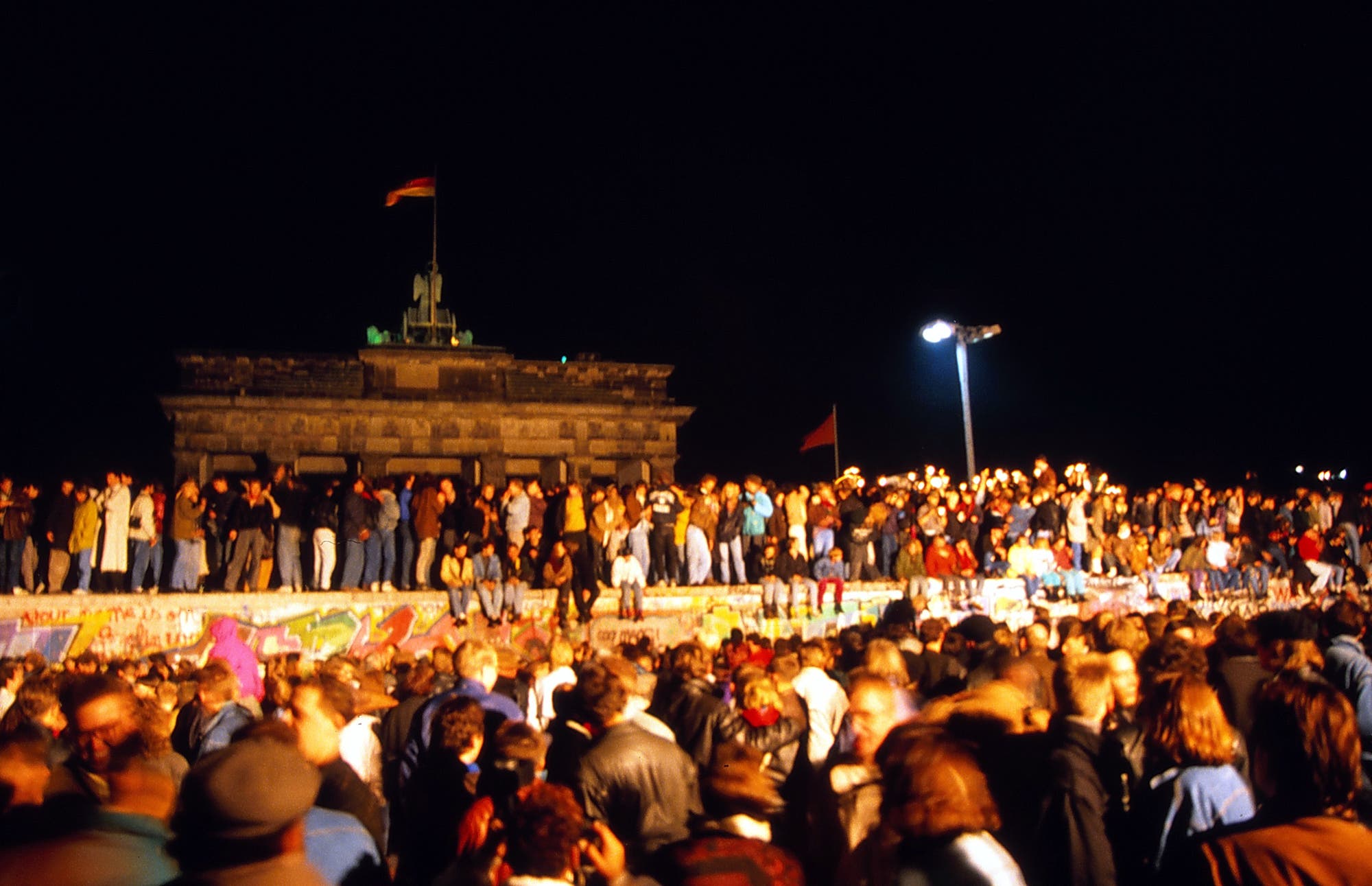 Menschen stehen und sitzen dicht an dicht auf der Berliner Mauer. Im Hintergrund ist das Brandenburger Tor zu erkennen. Im Vordergrund steht eine Volksmenge vor der Mauer.