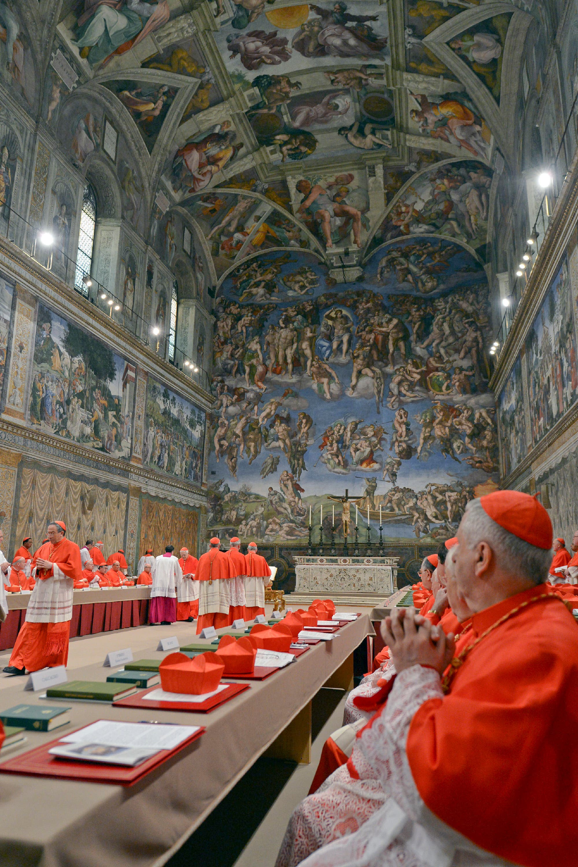 Eine Gruppe von Kardinälen in roten Gewändern versammelt sich in der Sixtinischen Kapelle, die mit den berühmten Fresken von Michelangelo verziert ist. Im Vordergrund sitzt ein Kardinal mit gefalteten Händen an einem Tisch, auf dem rote Birette und Dokumente liegen. Die Kapelle ist reich mit religiösen Kunstwerken dekoriert, die die Wände und die Decke bedecken. Die Szene vermittelt eine feierliche und bedeutungsvolle Atmosphäre.