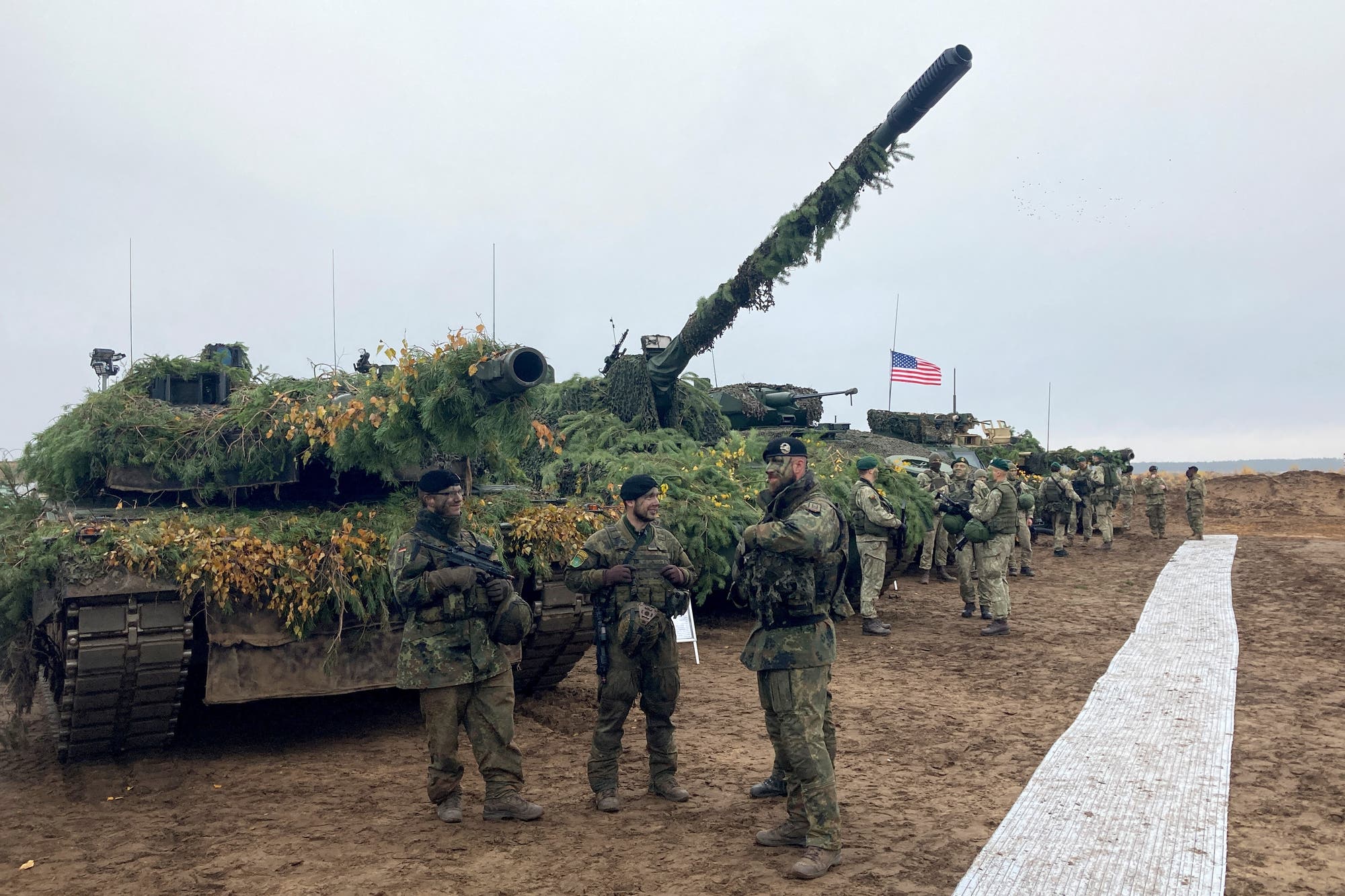 Mit Zweigen getarnte Militärfahrzeuge stehen in einer Reihe, davor unterhalten sich Soldaten