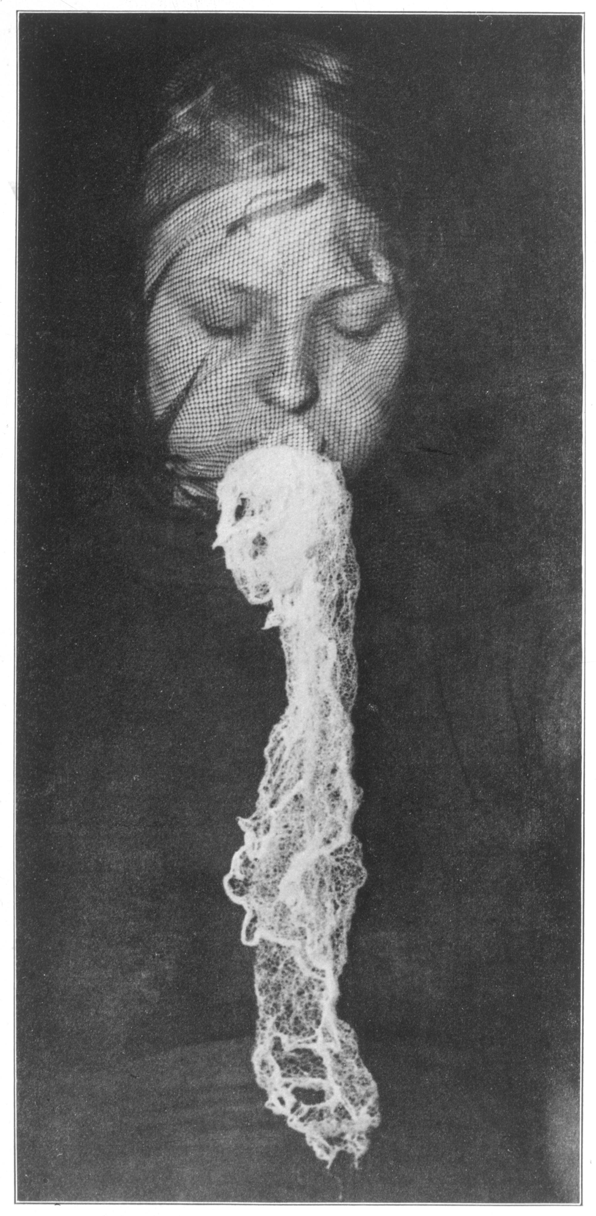 Eine schwarz gekleidete Person mit geschlossenen Augen scheint ein langes, helles, spitzenartiges Material aus dem Mund heraushängen zu haben. Die Szene auf dem Schwarz-Weiß-Bild wirkt mystisch und geheimnisvoll. 