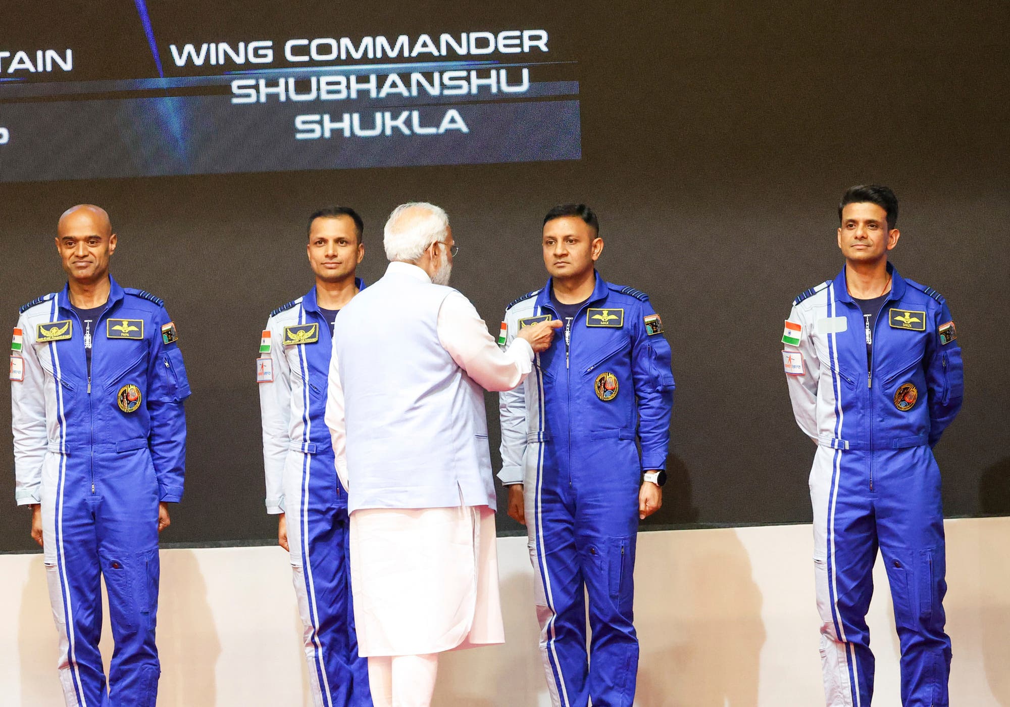 Eine Gruppe von vier Männern in blauen Raumanzügen steht auf einer Bühne. Ein Mann in traditioneller weißer Kleidung spricht mit ihnen. Auf der Leinwand im Hintergrund steht "Wing Commander Shubhanshu Shukla". Die Raumanzüge tragen Abzeichen und die indische Flagge.