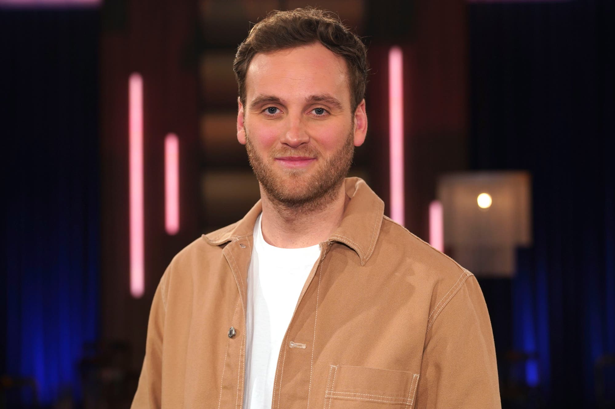 Ein Mann mit braunen Haaren trägt eine beige Jacke über einem weißen T-Shirt und steht vor einem unscharfen Hintergrund mit vertikalen Lichtern.