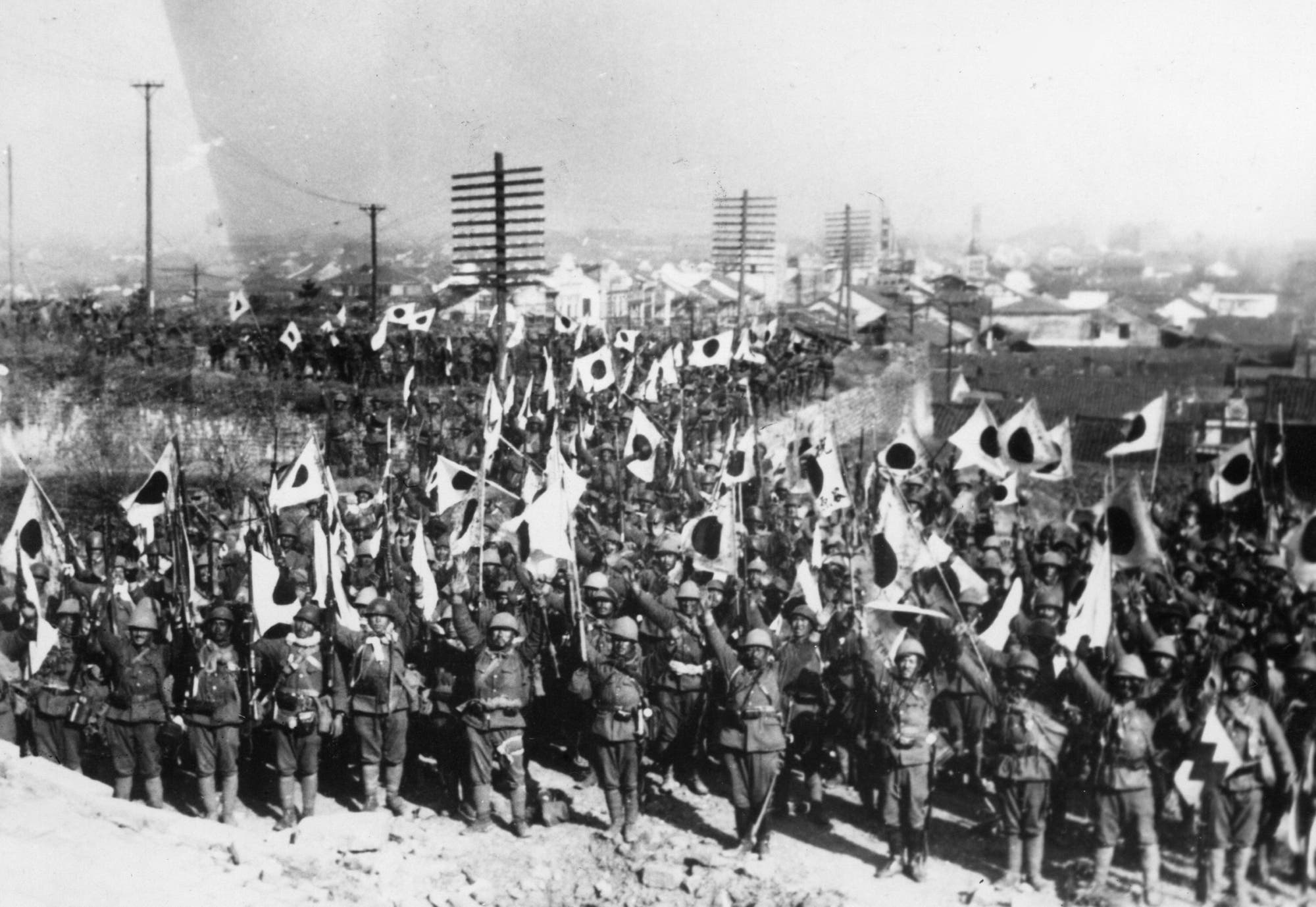Eine große Gruppe von Soldaten marschiert in Formation durch eine Stadt, während sie Fahnen mit kreisförmigen Symbolen schwenken. Im Hintergrund sind Gebäude und Strommasten zu sehen. Die Szene vermittelt eine militärische Parade oder einen Marsch.