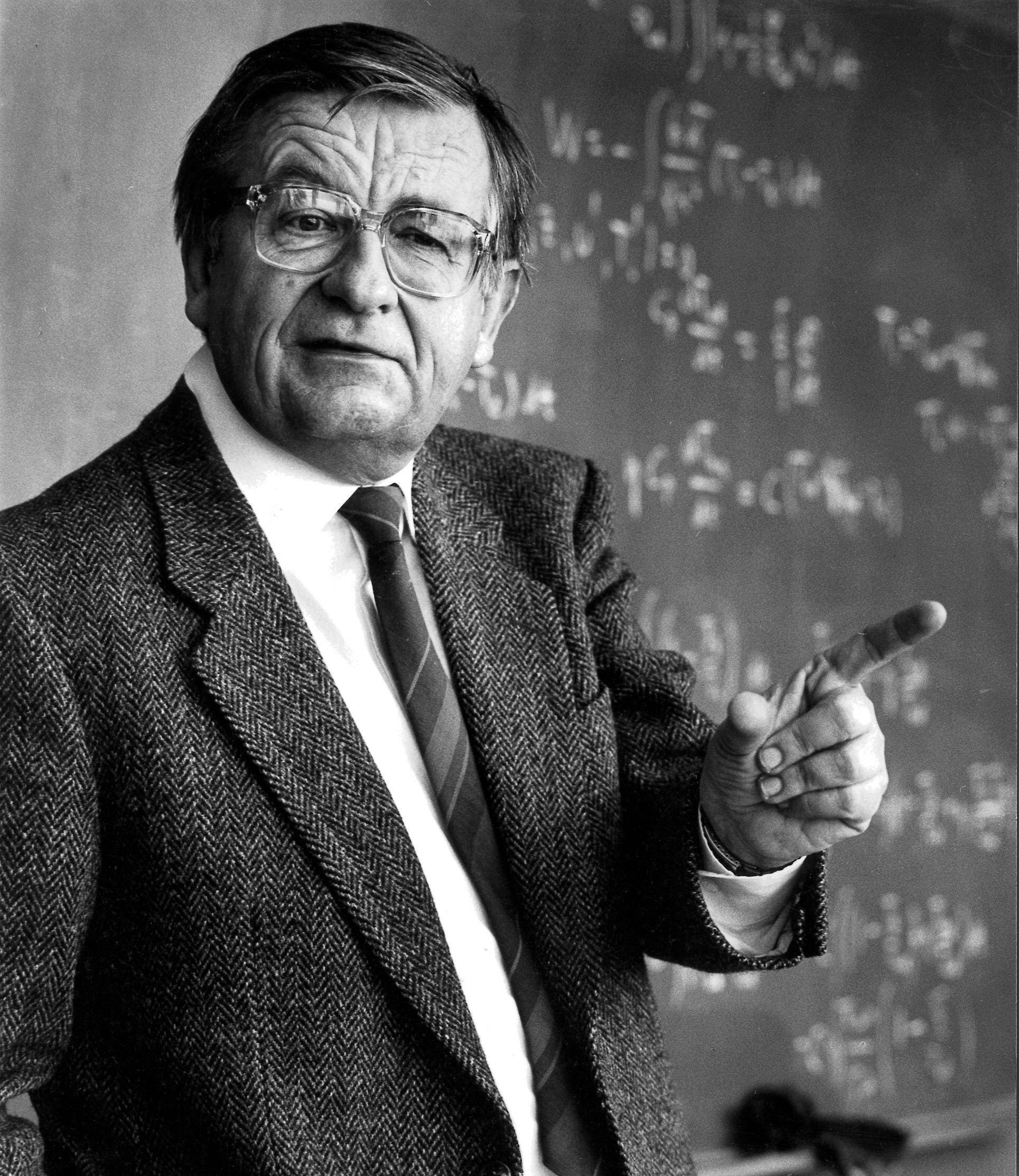 Ein älterer Mann mit grauem Haar und Brille trägt einen dunklen Anzug und eine gemusterte Krawatte. Er steht vor einem Hintergrund mit grünen Blättern und schaut ruhig in die Kamera.