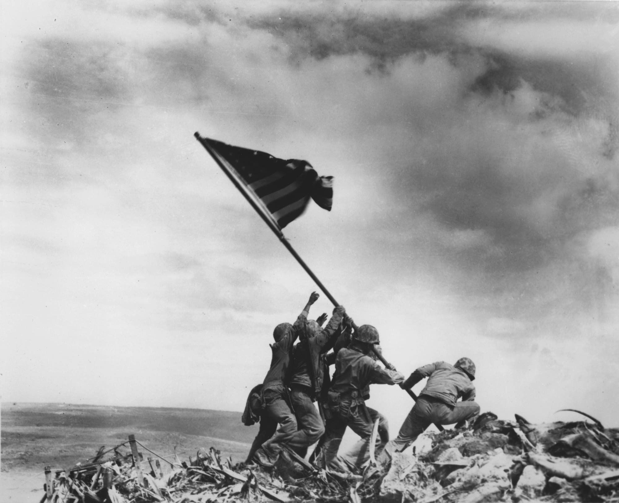 Auf einer Schwarz-weiß-Aufnahme sind fünf Soldaten zu sehen, die gemeinsam eine Fahne der Vereinigten Staaten in den Boden rammen. Die Flagge weht im Wind, der hell bewölkt ist.