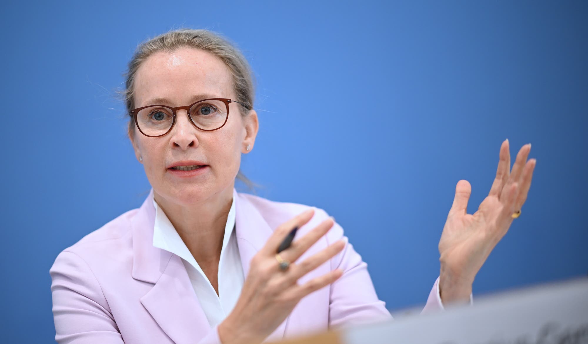 Eine Person in einem rosafarbenen Blazer und weißem Hemd spricht gestikulierend auf einer Pressekonferenz. Sie hat blondes Haar, trägt eine Brille und hält einen Stift in der Hand. Der Hintergrund ist blau. 