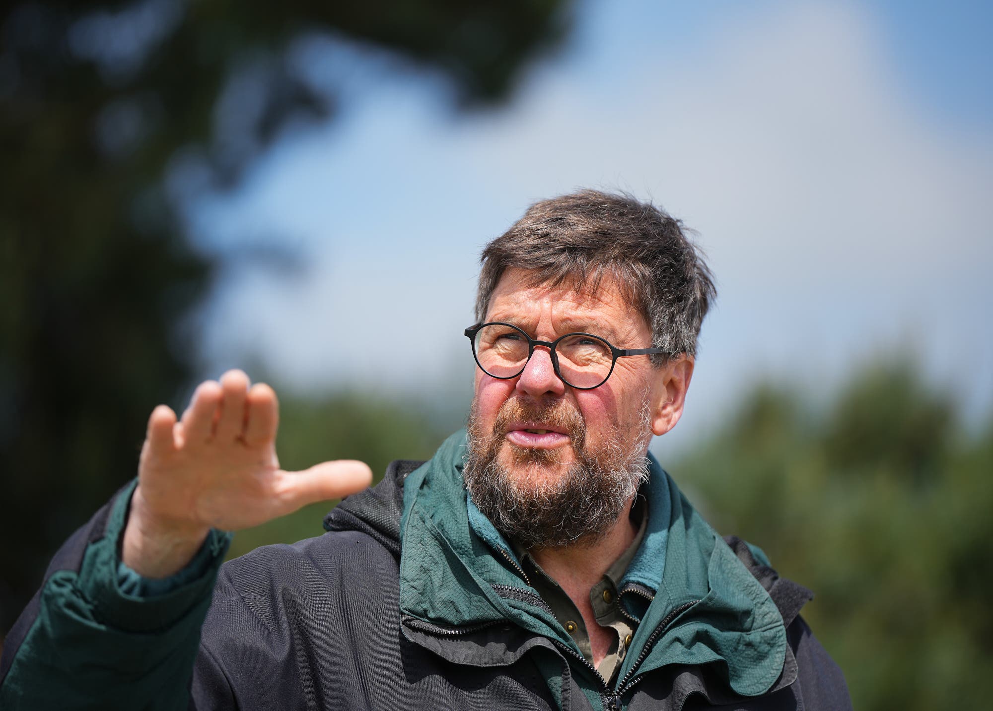 Ein Mann mit Brille und Bart steht im Freien und gestikuliert mit der rechten Hand. Er trägt eine dunkle Jacke und einen grünen Schal. Der Hintergrund ist unscharf, mit Bäumen und einem blauen Himmel.