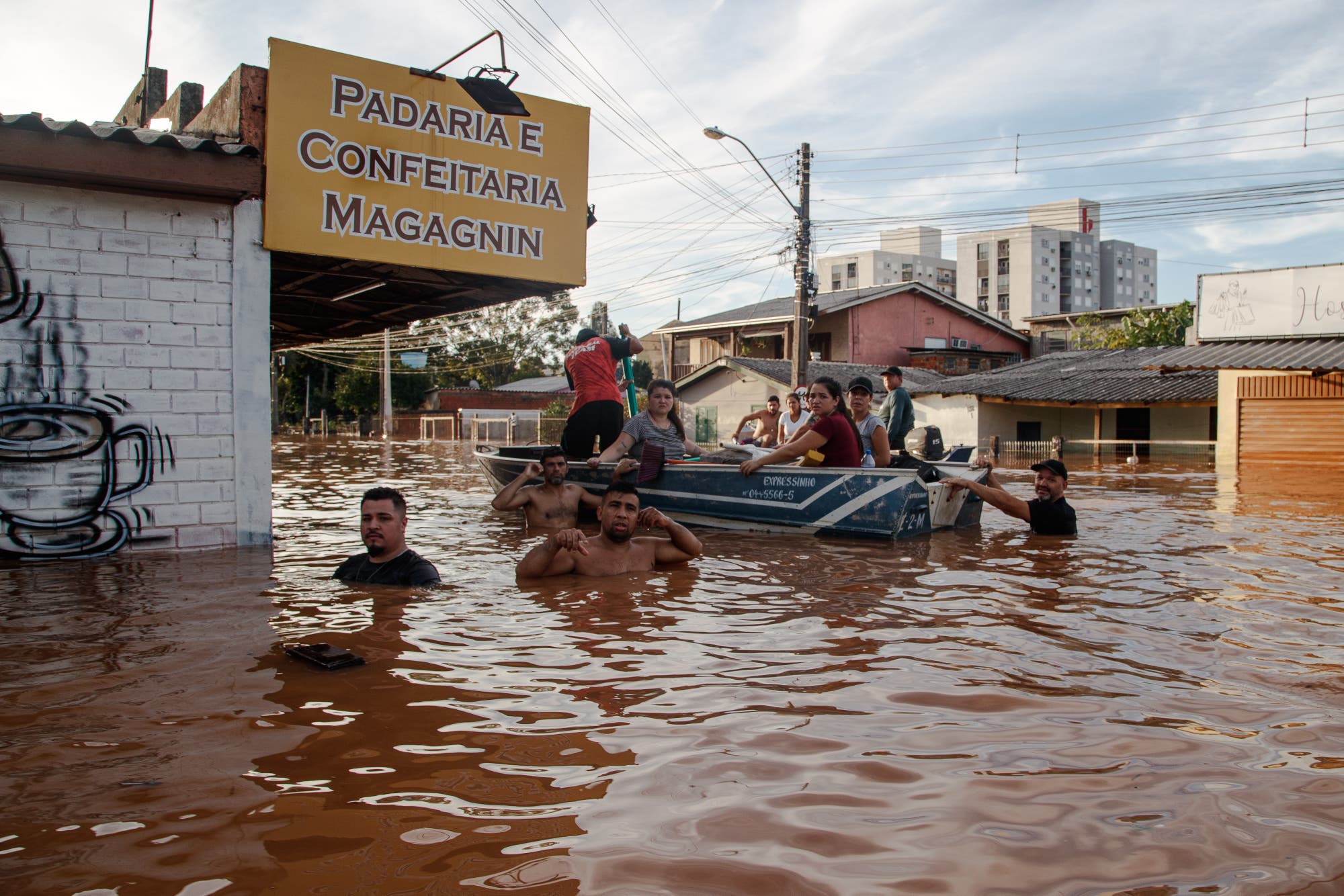Ein Rettungsteam aus mehreren Männern evakuiert Frauen, Kinder und Männer in einem Boot in Rio Grande do Sul. Die Retter stehen bis zur Brust im braunen Wasser vor einer Bäckerei mit einem gelben Werbeschild.