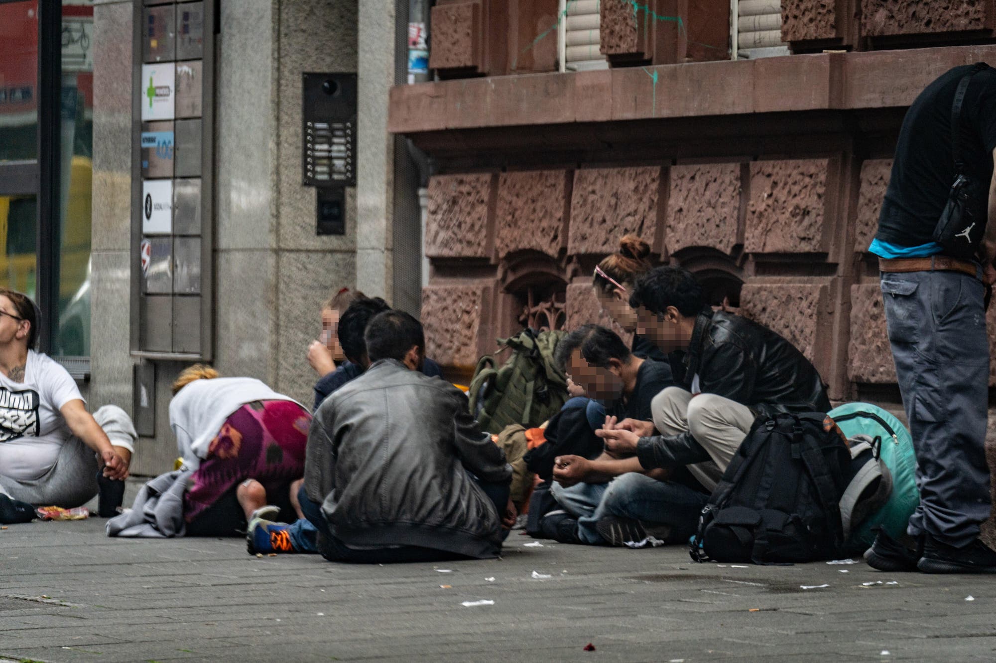 Eine Gruppe von Menschen sitzt auf dem ungepflegten Bürgersteig vor einem Gebäude mit einer roten Backsteinfassade. Einige sind dabei, ihren Drogenkonsum vorzubereiten und einige sind in Gespräche vertieft. Die Umgebung wirkt urban.