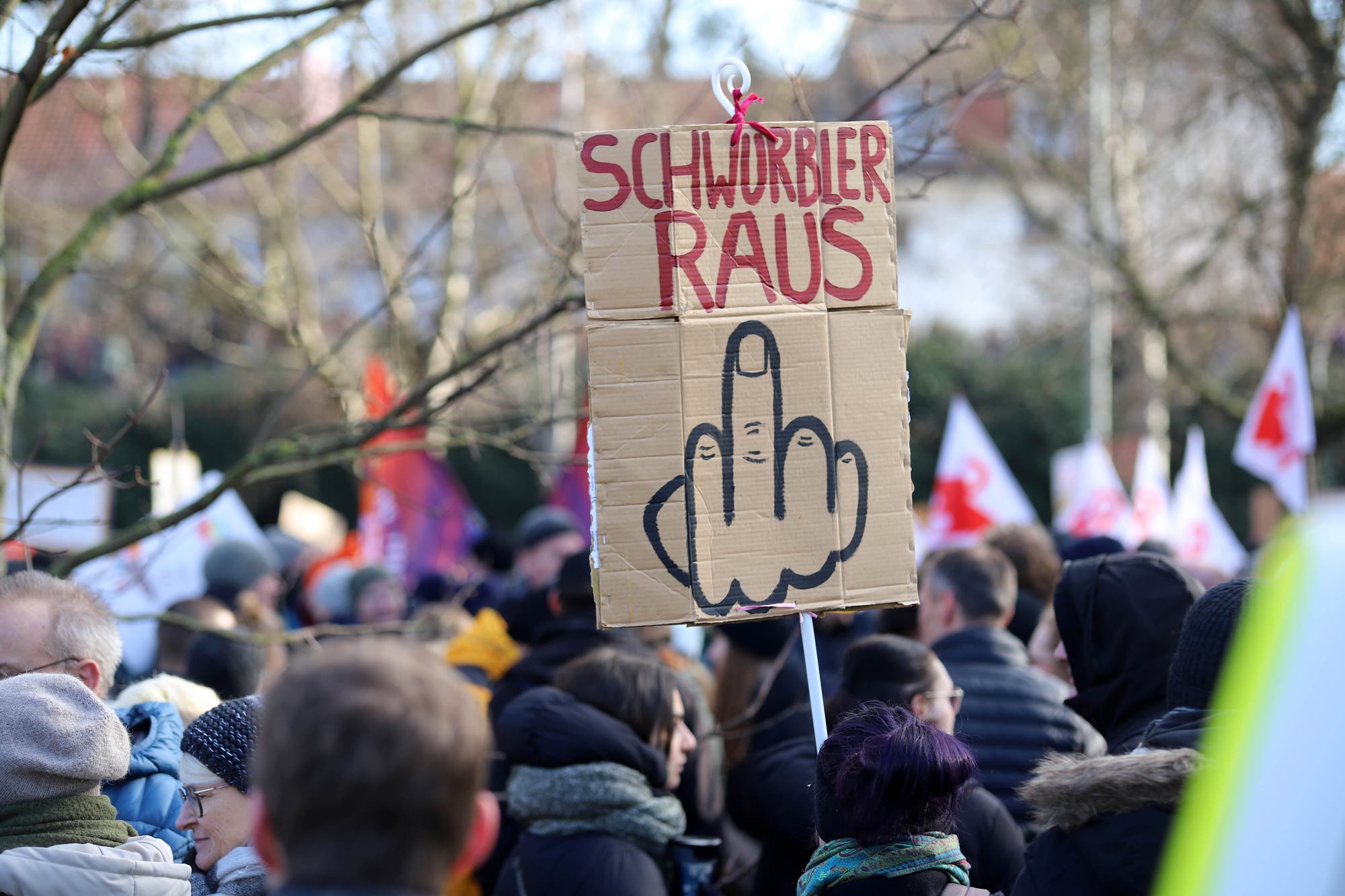 Eine Menschenmenge bei einer Demonstration im Freien. Im Vordergrund wird ein Pappschild hochgehalten, auf dem "Schwurbler raus" steht, begleitet von einer Zeichnung einer Hand mit ausgestrecktem Mittelfinger. Im Hintergrund sind weitere Demonstranten und Fahnen zu sehen. Die Szene vermittelt eine protestierende Stimmung.