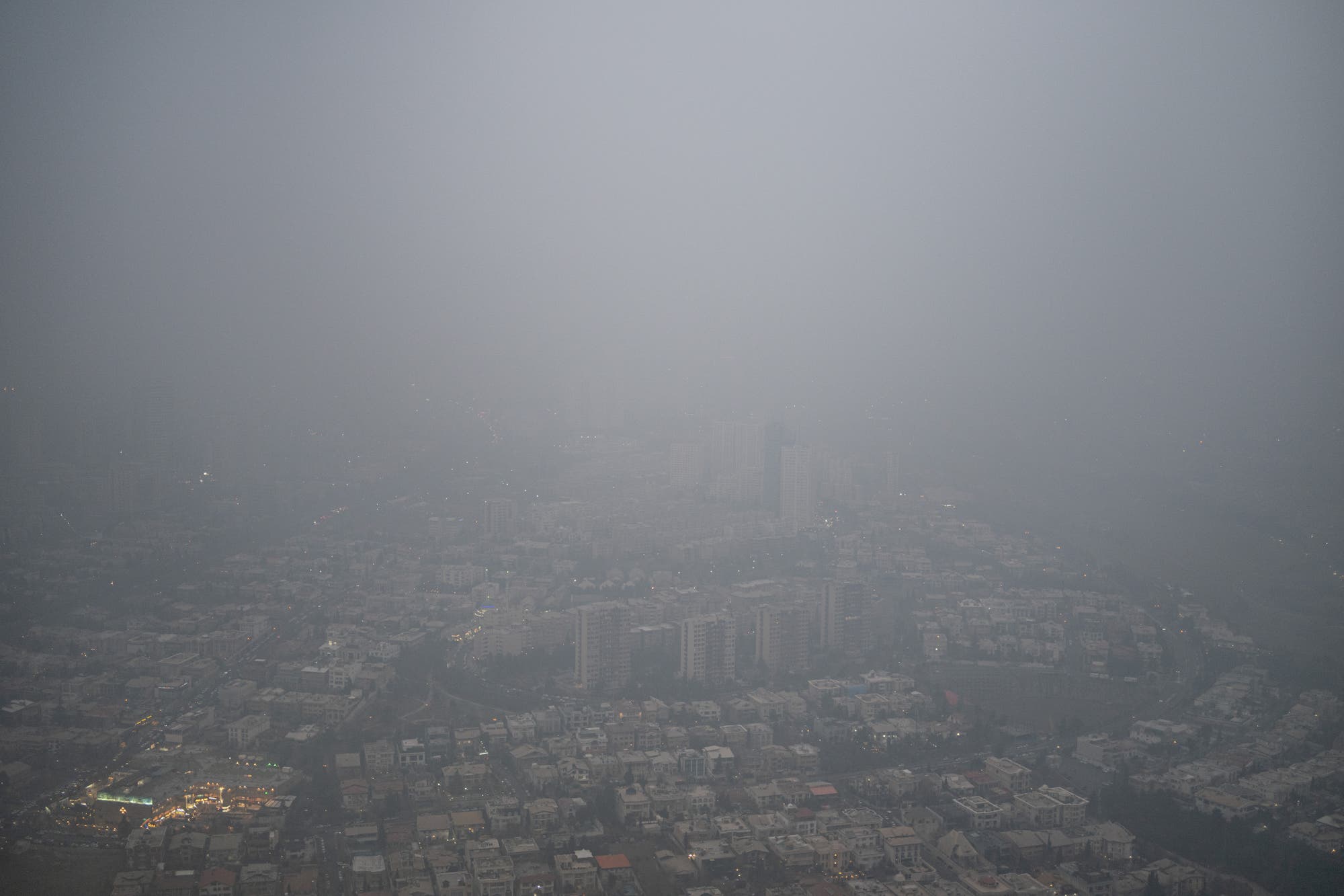 Luftaufnahme der Stadt Teheran, die in dichten Smog gehüllt ist. Die Sicht ist stark eingeschränkt, und die Gebäude sind nur schemenhaft zu erkennen. Die Beleuchtung der Stadt ist gedämpft, was auf eine hohe Luftverschmutzung hinweist. Keine Menschen sind sichtbar. Luftaufnahme der Stadt Teheran, die in dichten Smog gehüllt ist. Die Sicht ist stark eingeschränkt, und die Gebäude sind nur schemenhaft zu erkennen. Die Beleuchtung der Stadt ist gedämpft, was auf eine hohe Luftverschmutzung hinweist. Keine Menschen sind sichtbar.