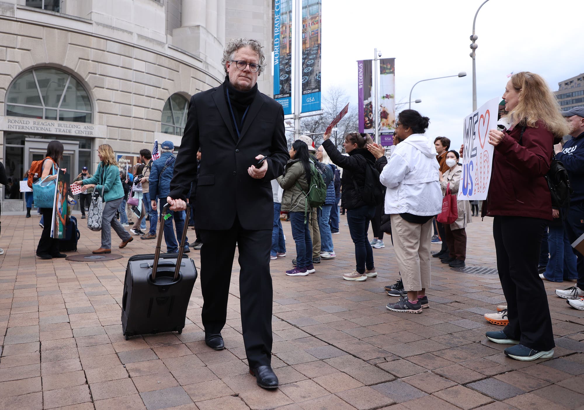 Ein Mann in einem schwarzen Anzug zieht einen Rollkoffer über einen gepflasterten Platz vor dem Ronald Reagan Building and International Trade Center. Im Hintergrund versammeln sich Menschen, einige halten Schilder hoch, darunter eines mit der Aufschrift "WE <3 US". Die Szene deutet auf eine Versammlung oder Demonstration hin. Die Umgebung ist urban, mit großen Gebäuden und Fahnenmasten.