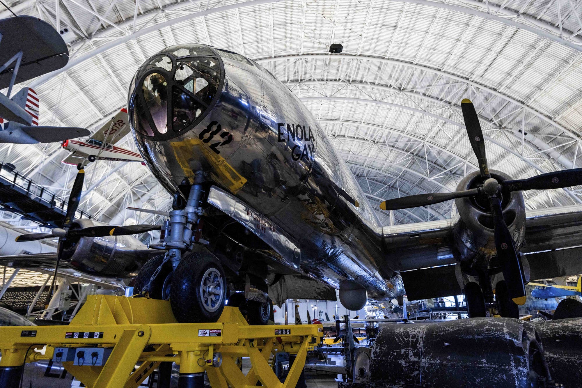 Ein silbernes Flugzeug, die »Enola Gay«, ausgestellt in einem großen, gewölbten Hangar. Das Flugzeug steht auf gelben Stützen, die seine Räder tragen. Im Hintergrund sind weitere Flugzeuge zu sehen. Die Umgebung ist gut beleuchtet und zeigt die Struktur des Hangars.