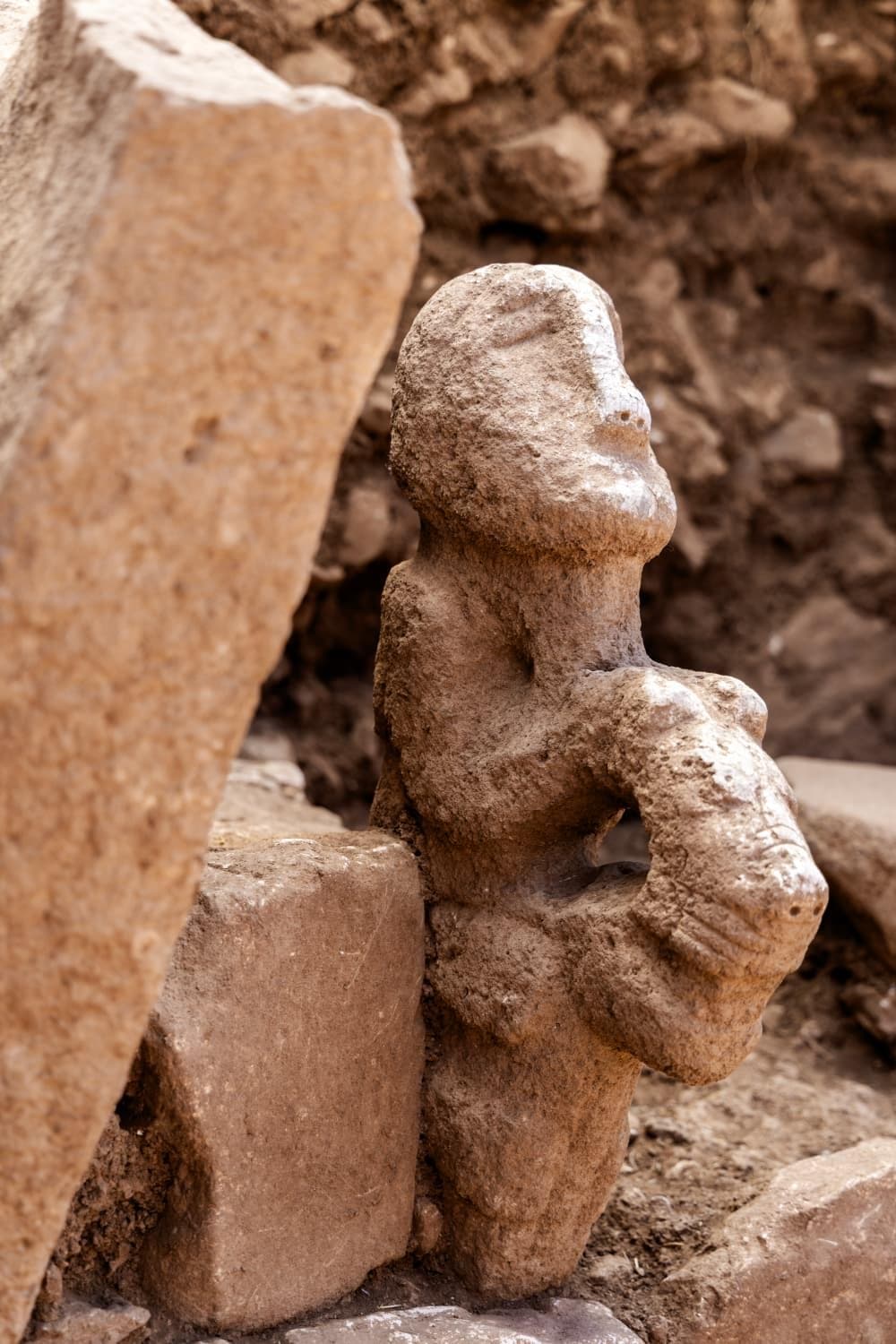 Eine grob behauene Steinskulptur, die eine menschliche Figur darstellt, steht zwischen großen, unregelmäßigen Steinen in einer archäologischen Ausgrabungsstätte. Die Figur hat keine klaren Gesichtszüge und hält ein undefinierbares Objekt. Der Hintergrund zeigt erdige, steinige Strukturen, die auf eine historische oder prähistorische Umgebung hindeuten.