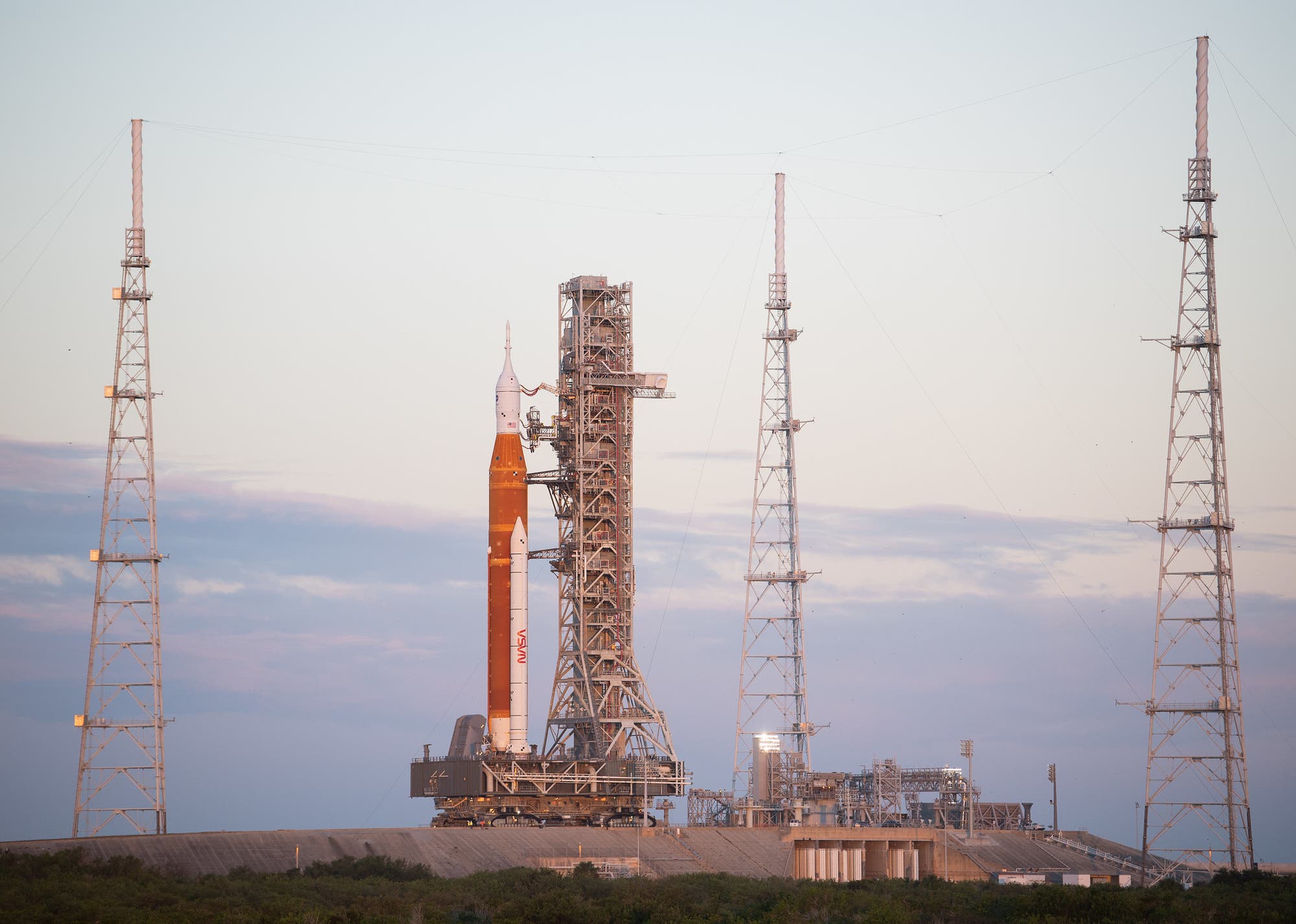 Eine große Rakete steht auf einer Startrampe, umgeben von mehreren hohen Türmen. Die Rakete ist orange-weiß und trägt das NASA-Logo. Der Himmel im Hintergrund ist leicht bewölkt und die Szene wird von sanftem Tageslicht beleuchtet. Die Startrampe ist von grüner Vegetation umgeben.