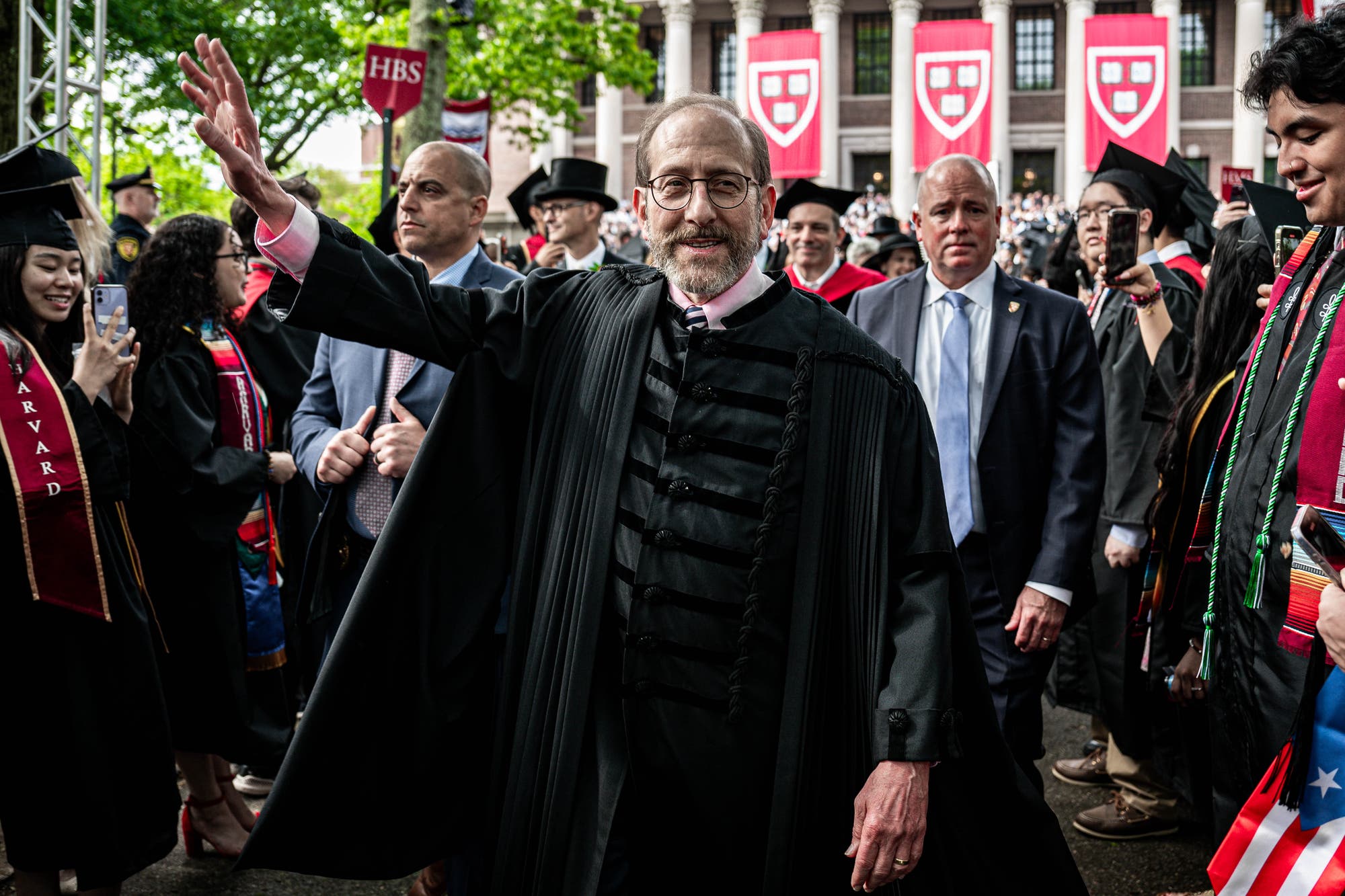 Eine Person in einem traditionellen akademischen Gewand winkt während einer Abschlussfeier auf dem Campus der Harvard University. Im Hintergrund sind Absolventen in Roben und Hüten zu sehen, einige machen Fotos. Rote Banner mit dem Harvard-Logo hängen an einem Gebäude. Die Atmosphäre ist feierlich und festlich.