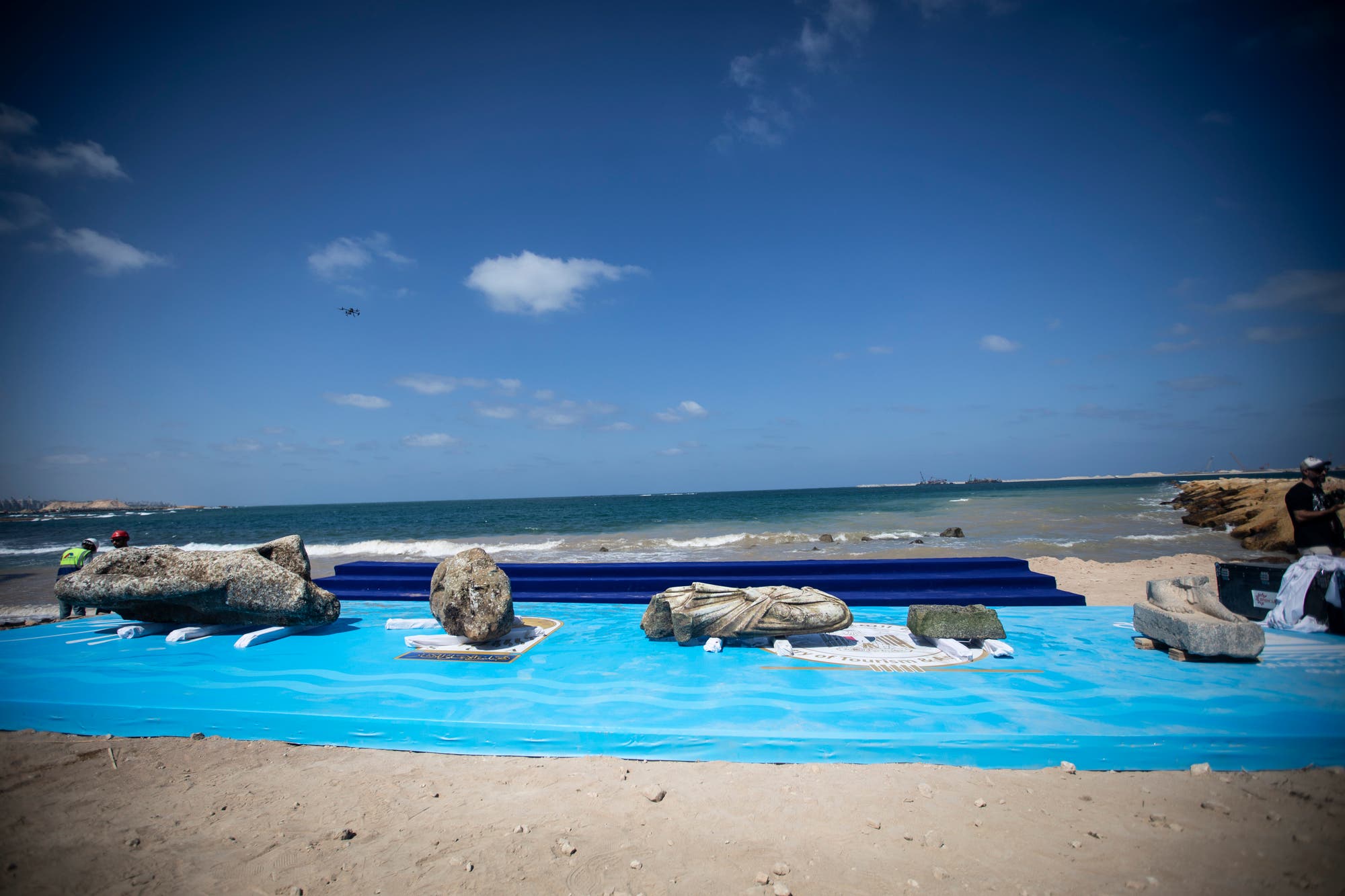 Eine Strandlandschaft mit mehreren antiken Steinskulpturen, die auf blauen Plattformen am Ufer ausgestellt sind. Im Hintergrund ist das Meer mit leichtem Wellengang zu sehen, darüber ein klarer Himmel mit wenigen Wolken. Links im Bild ist eine Person mit Schutzhelm zu erkennen, die sich den Skulpturen nähert. Rechts steht eine weitere Person neben den Exponaten.