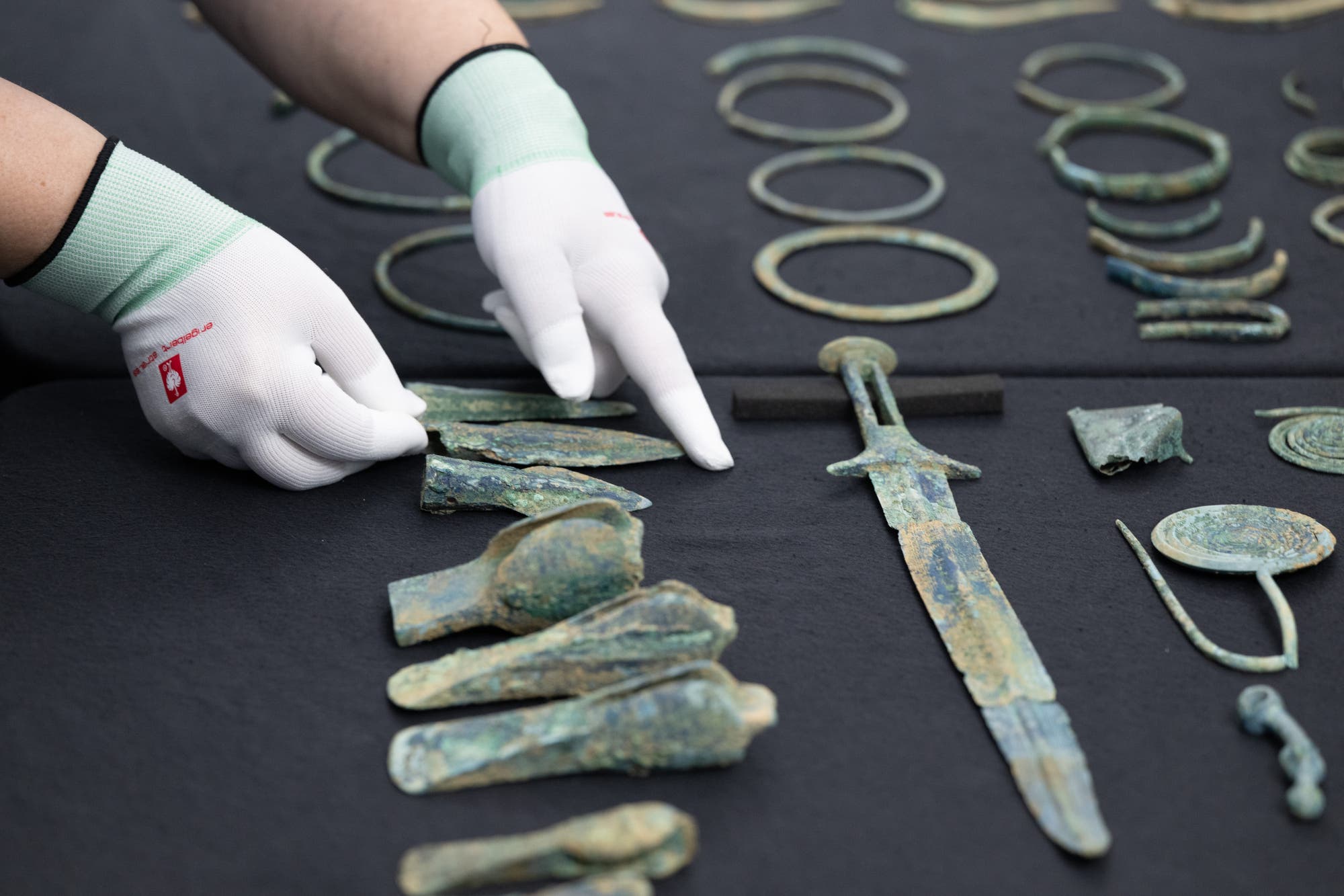 Eine Person mit weißen Handschuhen zeigt auf eine Sammlung alter Bronzeobjekte, die auf einem dunklen Tuch ausgebreitet sind. Zu den Artefakten gehören ein Schwert, Ringe und verschiedene Werkzeuge. Die Szene deutet auf eine archäologische Untersuchung oder Präsentation hin. Eine Person mit weißen Handschuhen zeigt auf eine Sammlung alter Bronzeobjekte, die auf einem dunklen Tuch ausgebreitet sind. Zu den Artefakten gehören ein Schwert, Ringe und verschiedene Werkzeuge. Die Szene deutet auf eine archäologische Untersuchung oder Präsentation hin.
