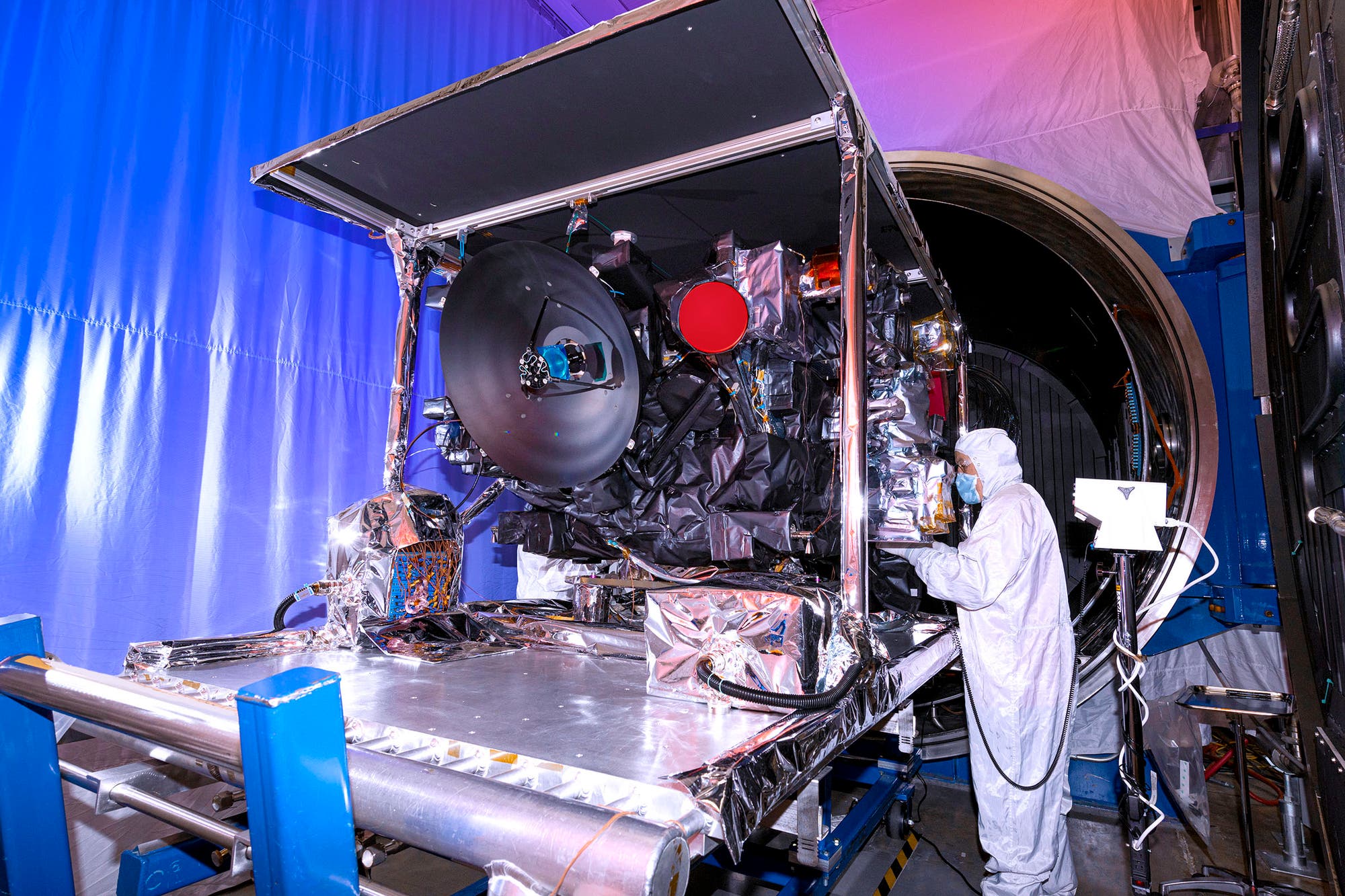 Eine Person in einem Reinraum-Anzug arbeitet an einem großen, mit Folie bedeckten wissenschaftlichen Gerät in einem Labor. Das Gerät hat eine große, runde Antenne und ist von verschiedenen technischen Komponenten umgeben. Der Raum ist in blaues Licht getaucht, im Hintergrund ist eine große, runde Öffnung zu sehen.