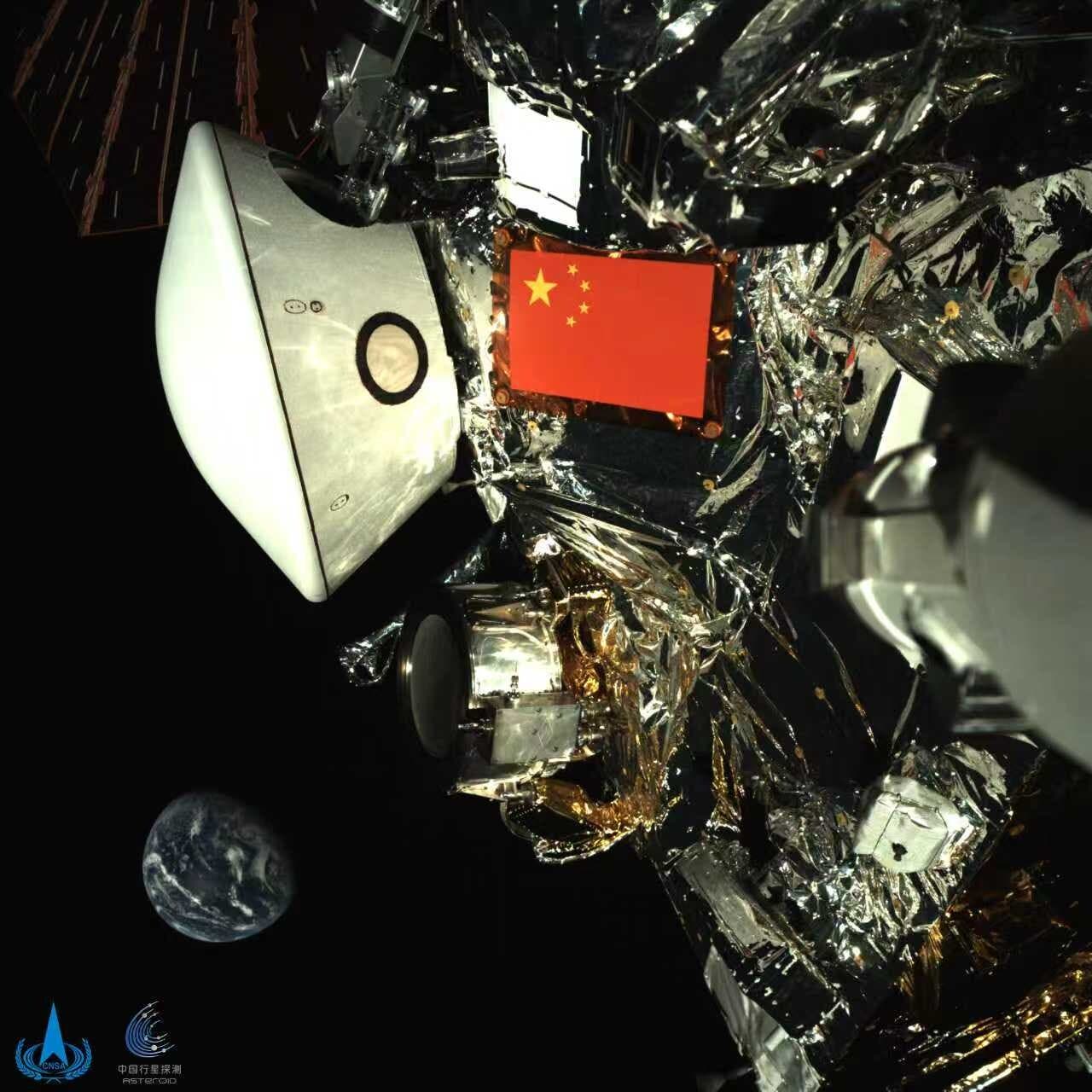 Eine Raumsonde im Weltraum, umgeben von reflektierenden Materialien und technischen Komponenten. Eine chinesische Flagge ist prominent auf der Sonde zu sehen. Im Hintergrund ist die Erde als kleiner blauer Planet sichtbar. Das Bild zeigt die Komplexität und den technologischen Fortschritt der Raumfahrt. Unten links befinden sich Logos, die auf chinesische Raumfahrtorganisationen hinweisen.
