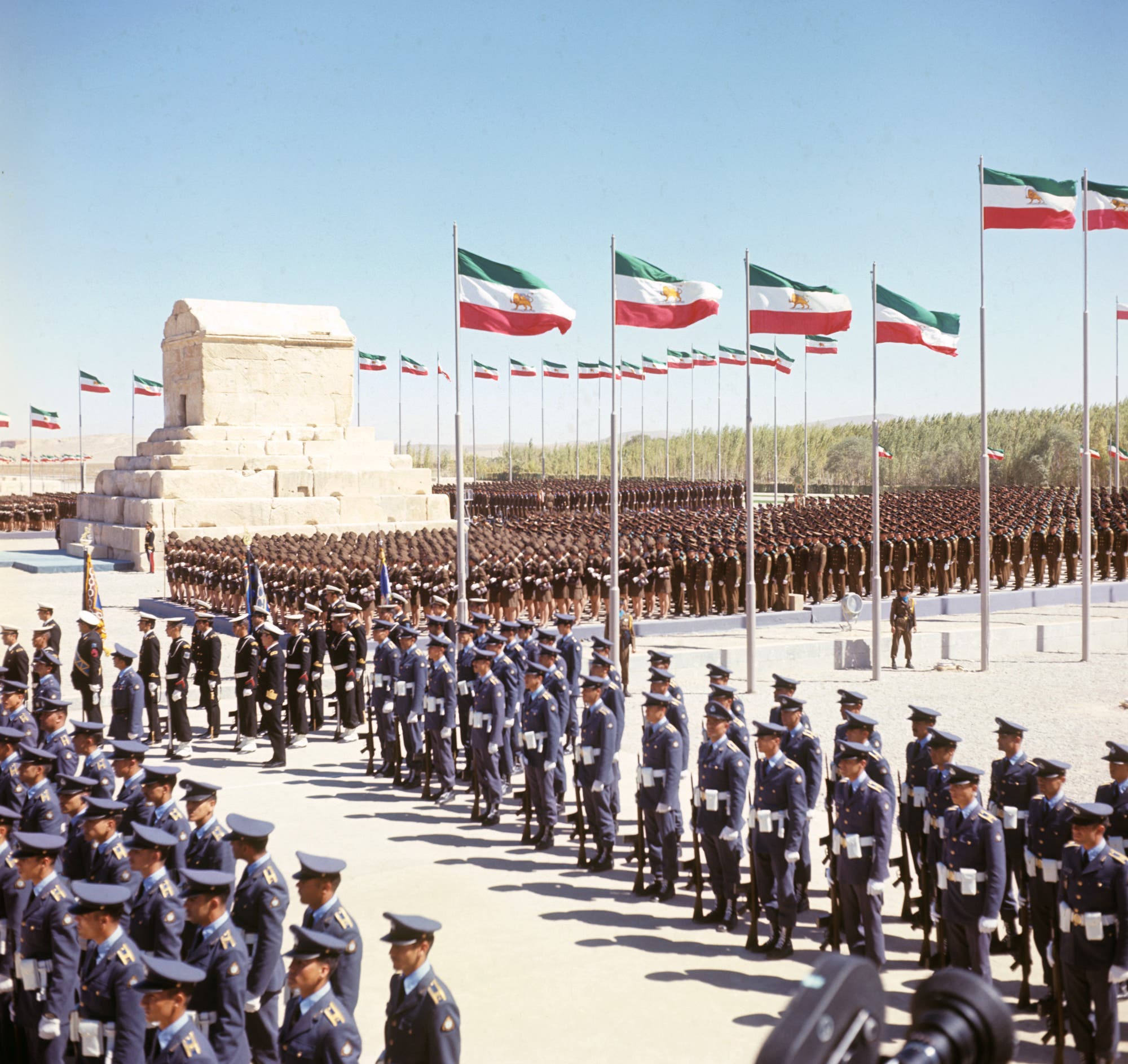 Eine große militärische Zeremonie mit Soldaten in blauen Uniformen, die in geordneten Reihen stehen. Im Hintergrund ist ein antikes Steingebäude zu sehen, umgeben von zahlreichen wehenden Flaggen. Die Szene findet unter klarem Himmel statt, und die Soldaten stehen stramm, während sie an der Zeremonie teilnehmen.