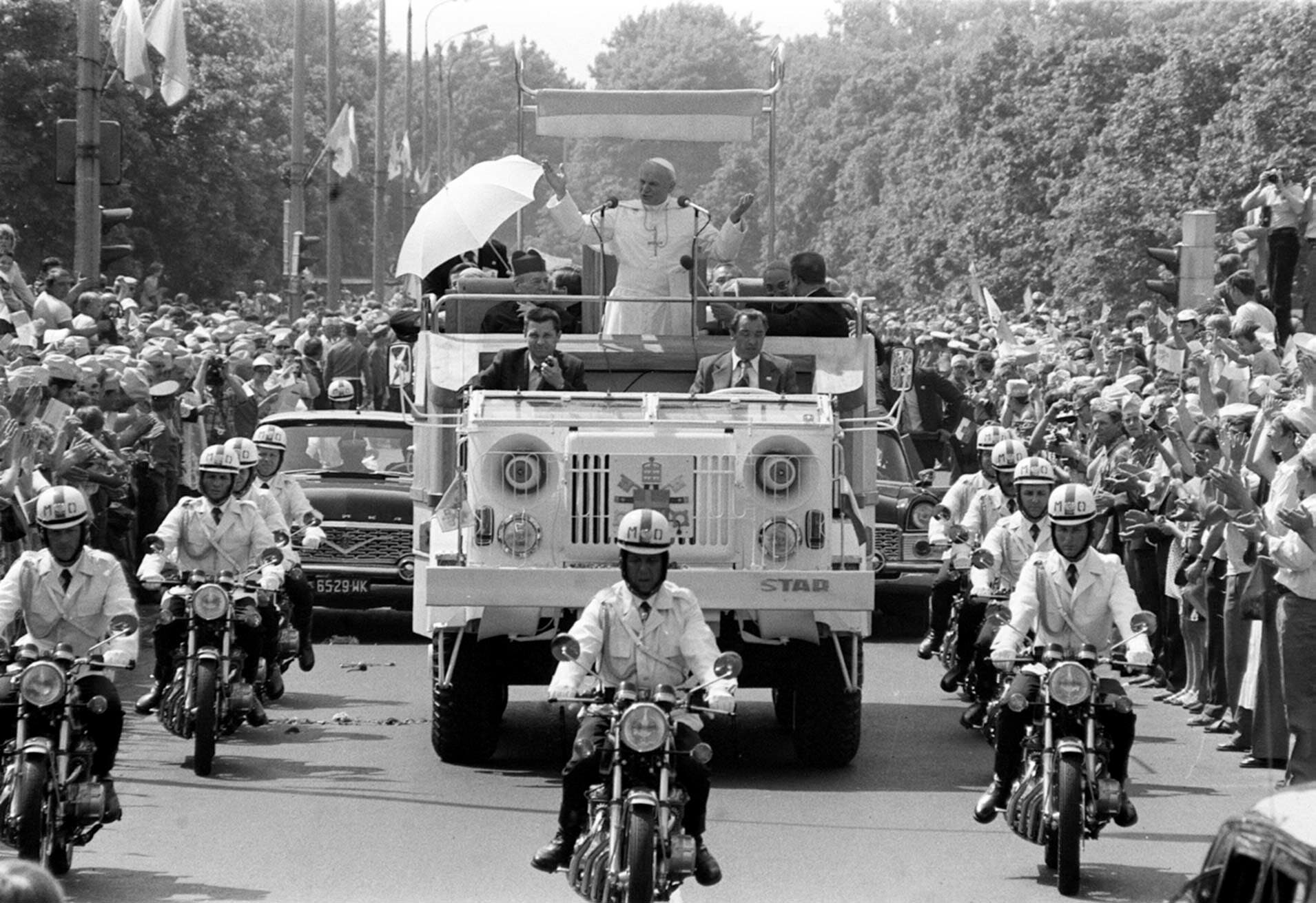 Das Schwarz-weiß-Foto zeigt Papst Johannes Paul II. stehend in einem weißen Auto, das eskortiert von Polizeimotorrädern über eine Straße fährt. An den Straßenrändern stehen sehr viele Menschen, um den Papst zu sehen.