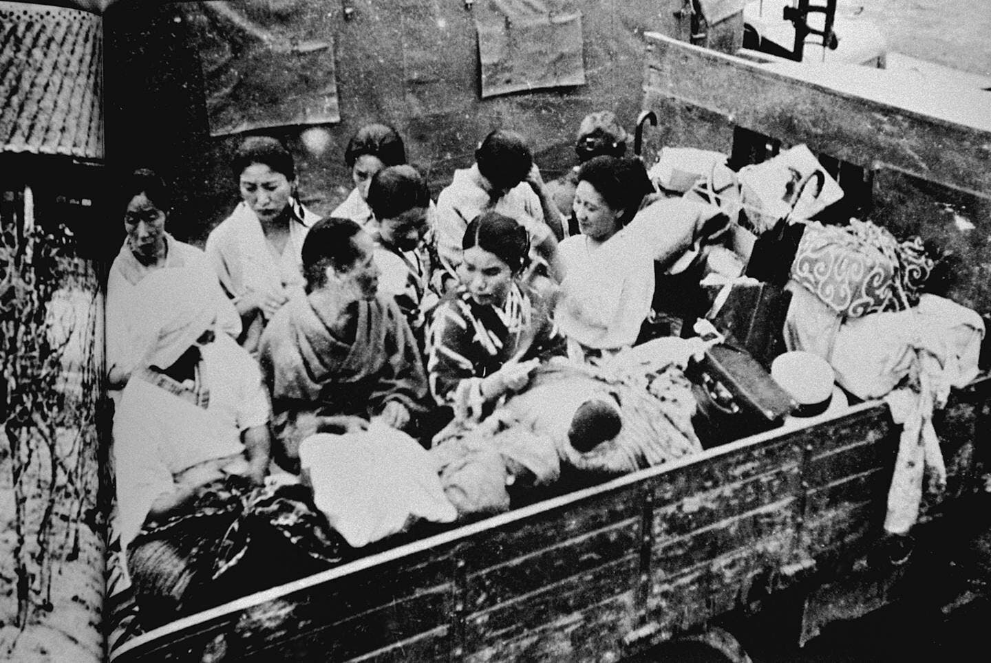 Eine Gruppe von Menschen sitzt dicht gedrängt auf der Ladefläche eines Lastwagens, umgeben von persönlichen Gegenständen und Gepäck. Die Szene wirkt angespannt und ernst, während die Personen miteinander interagieren. Der Lastwagen ist in einer städtischen Umgebung geparkt.