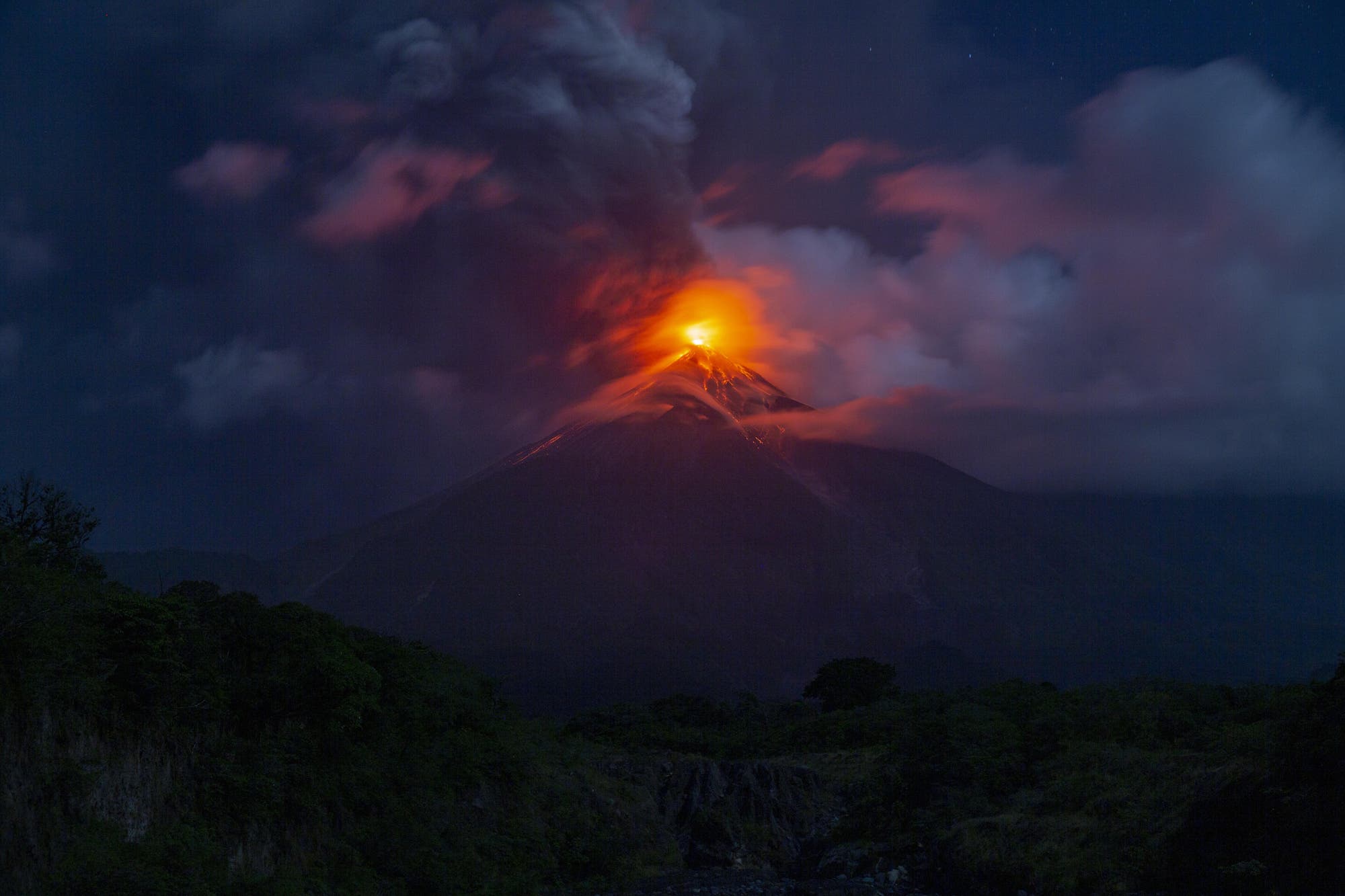 Ein Vulkan bricht bei Nacht aus und st&ouml;&szlig;t gl&uuml;hende Lava und dichten Rauch in den dunklen Himmel. Der leuchtend orangefarbene Krater hebt sich dramatisch von den dunklen Wolken und der umliegenden Landschaft ab. Im Vordergrund sind Silhouetten von B&auml;umen und H&uuml;geln zu erkennen.