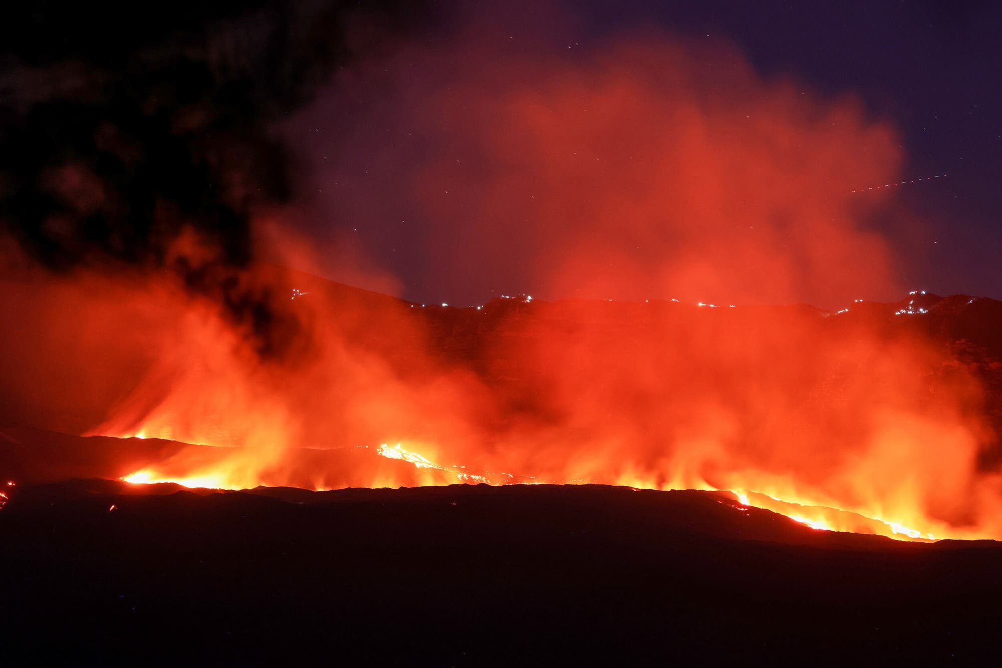 Ein nächtliches Foto eines Vulkanausbruchs, bei dem glühende Lava aus dem Krater fließt und orangefarbene Rauchwolken in den dunklen Himmel aufsteigen. Im Hintergrund sind schwach beleuchtete Hügel zu erkennen. Der Kontrast zwischen der hellen Lava und dem dunklen Himmel betont die Intensität des Ausbruchs. Ein nächtliches Foto eines Vulkanausbruchs, bei dem glühende Lava aus dem Krater fließt und orangefarbene Rauchwolken in den dunklen Himmel aufsteigen. Im Hintergrund sind schwach beleuchtete Hügel zu erkennen. Der Kontrast zwischen der hellen Lava und dem dunklen Himmel betont die Intensität des Ausbruchs.