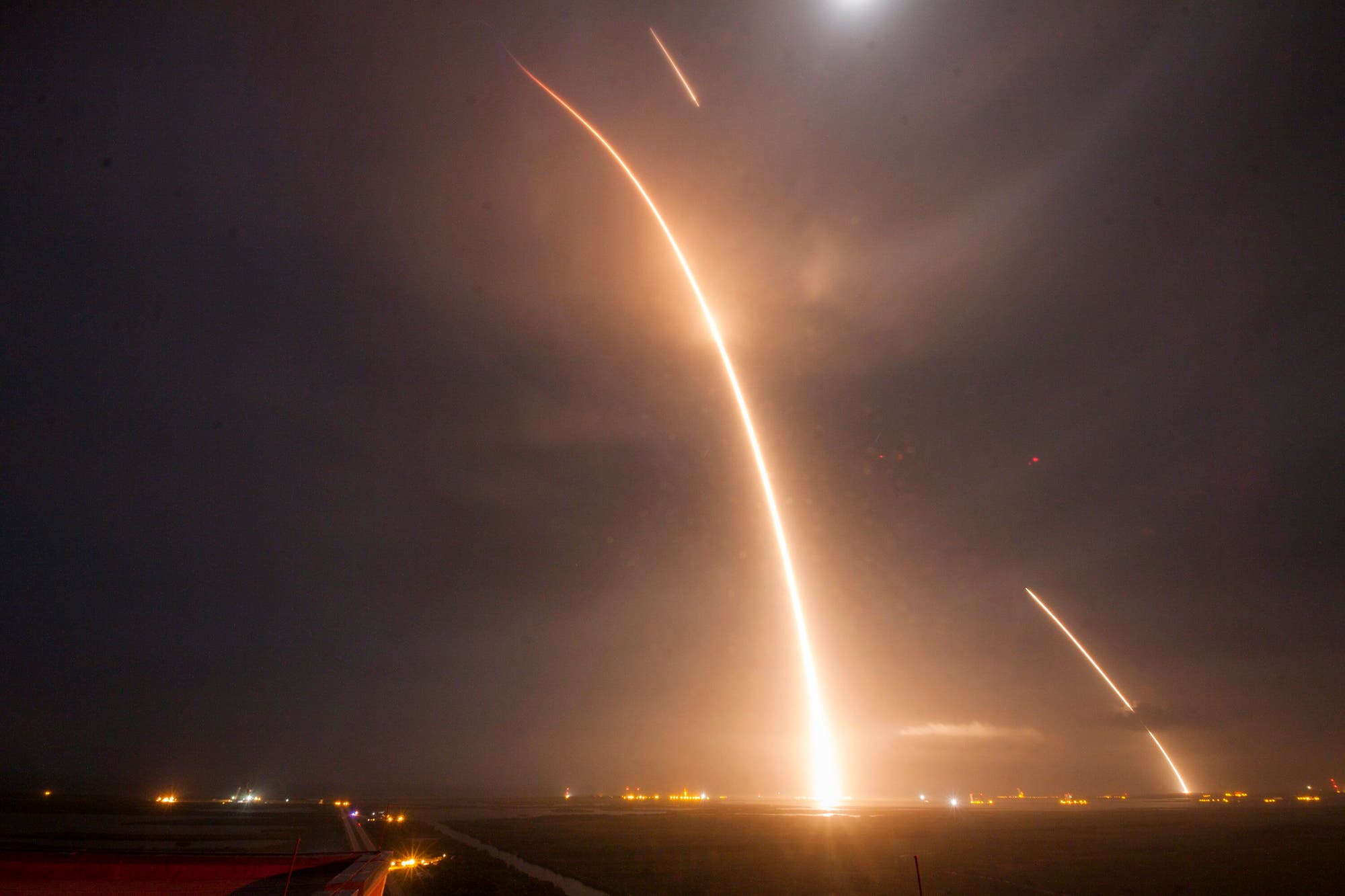 Eine Rakete startet in den Nachthimmel und hinterlässt eine leuchtende Spur. Der Himmel ist bewölkt, und am Horizont sind Lichter von Gebäuden oder Anlagen zu sehen. Die Szene vermittelt den Eindruck von Geschwindigkeit und technologischem Fortschritt.